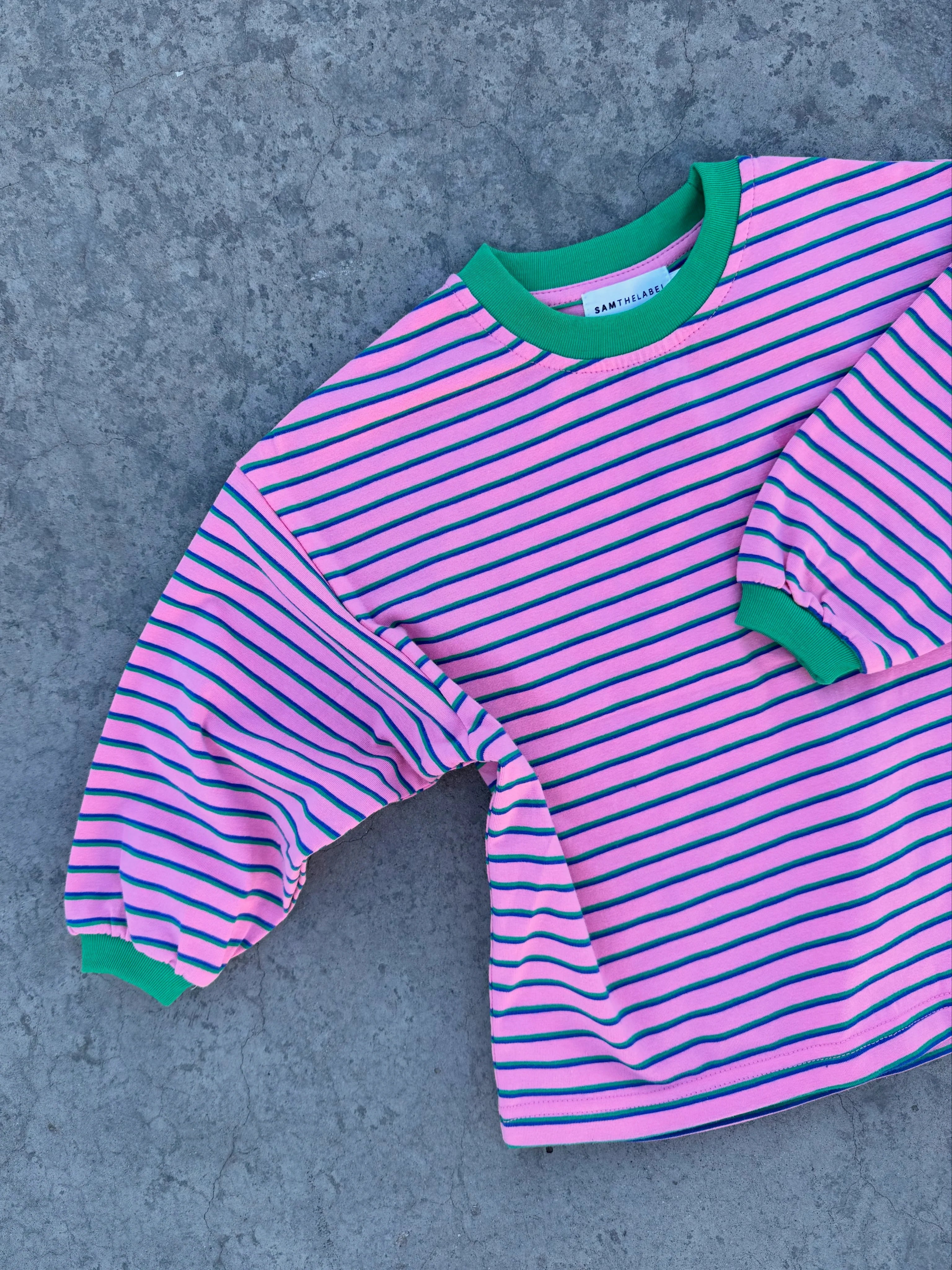 The Sam the Label - Kids Watermelon Sugar LS Tee by SAM THE LABEL, a unisex oversized shirt with pink and black stripes plus green cuffs and collar, is displayed on a textured gray surface.