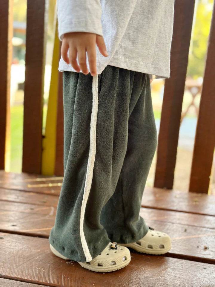 A child stands on a wooden deck wearing TINY TROVE's Darcy Racer Pants in Moss, featuring an elastic waistband and white side stripes, paired with a white long-sleeve shirt and beige slip-on shoes. The background is blurred, hinting at an outdoor setting with trees and a fence.