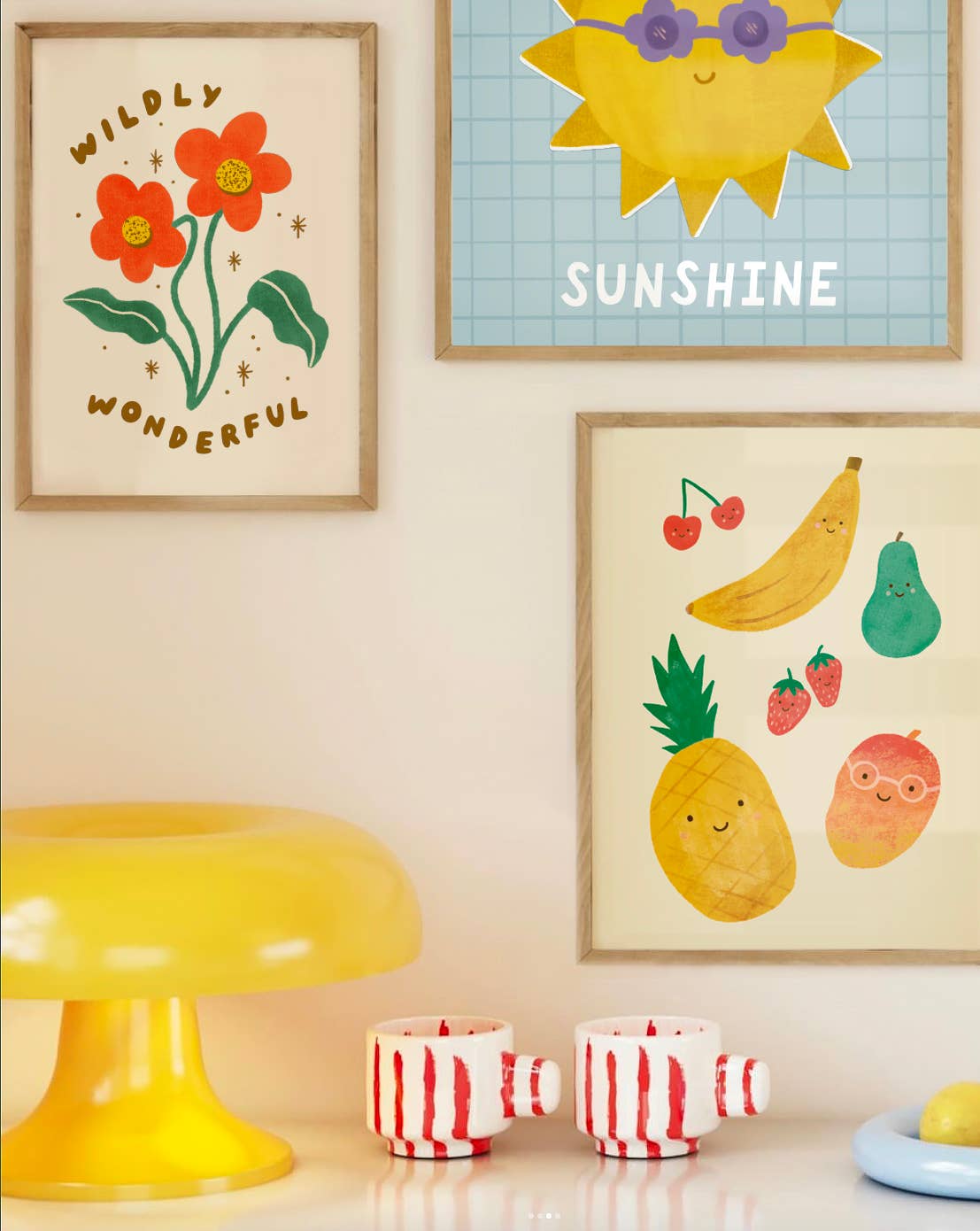 A picture of three art prints framed on a wall. One has the words wildly wonderful on it with two red flowers. The other has some cherries, a banana, strawberries, pear, pineapple and mango on it. The third picture is of a print with a sun wearing purple flower sunglasses with the words "you are my" above it and the word "sunshine" below, on a blue background with a square pattern on it. There is also a mushroom shaped art piece and two mugs with red stripes on them.