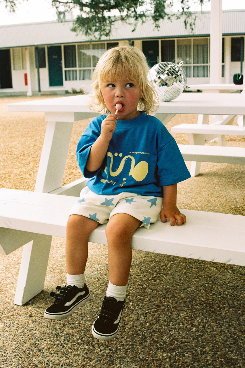 A young blonde child sits on a white wooden bench, licking a lollipop, wearing a Sunday Siblings Il Sole Tee in blue/yellow with star-patterned shorts, white socks, and black sneakers. A disco ball gleams on the bench behind them.