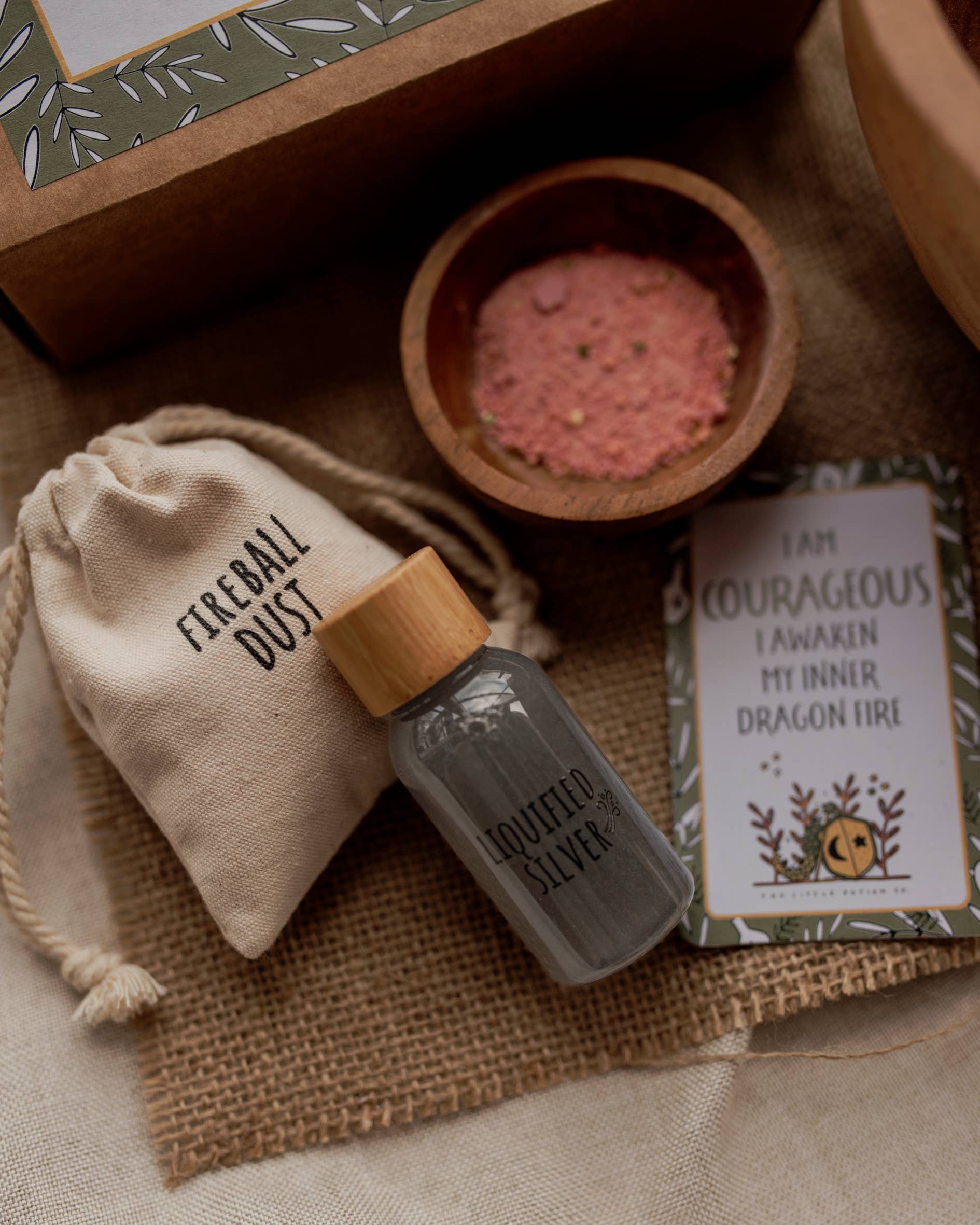 A flat-lay image features a small pouch labeled "Fireball Dust," a small bottle labeled "Liquefied Silver," a bowl of pink powder, and a card that reads "I am courageous, I awaken my inner dragon fire" with a leafy border and dragon illustration, showcasing the MINI Dragon Tales Potion Kit (A potion for courage) by THE LITTLE POTION CO—an imaginative play set perfect for aspiring dragons and knights.