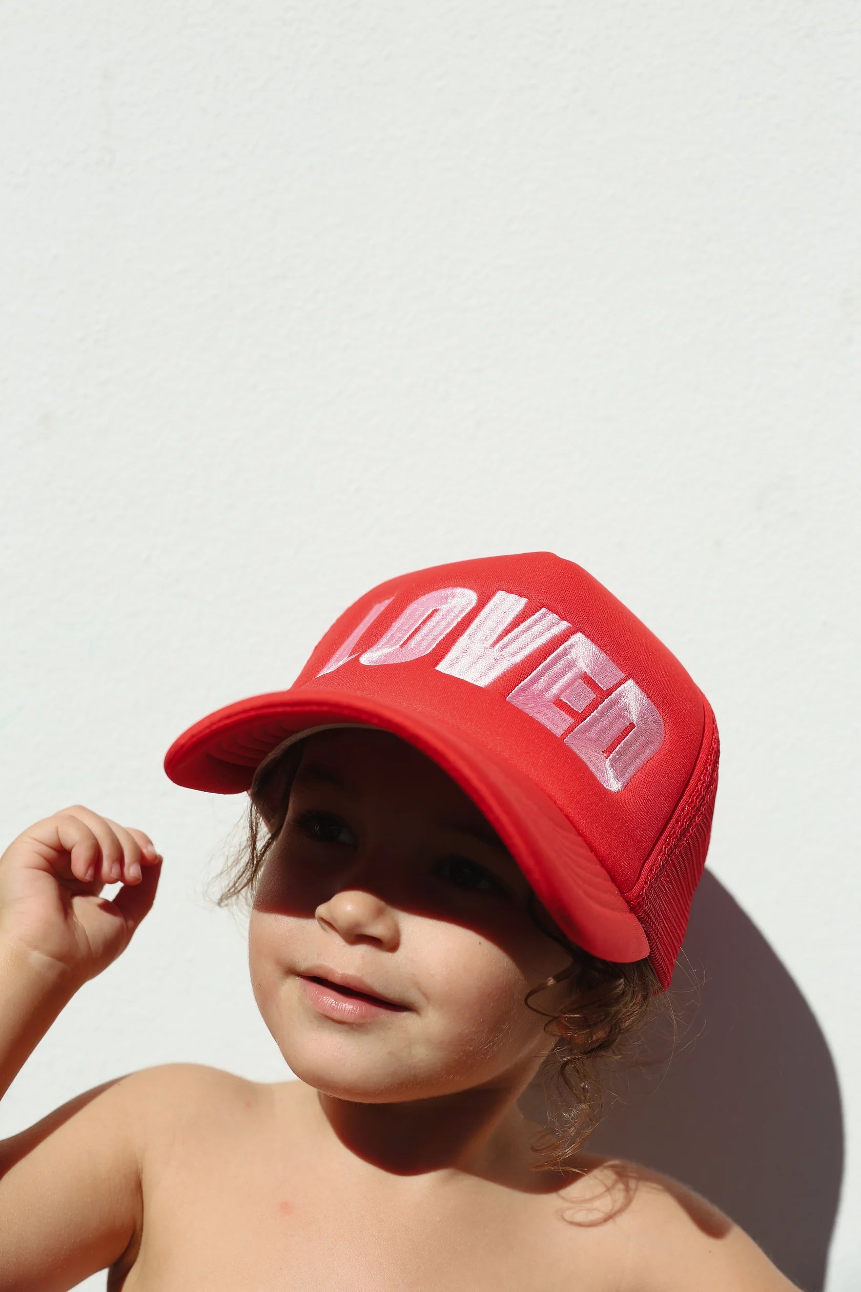 A young child with brown hair is wearing the "So Loved Trucker - Red" cap from TINY LOVE CLUB, which has the word "LOVED" embroidered in white. The child is shirtless and poses against a plain, light-colored background with a slight smile on their face, hand lifted towards the adjustable back strap.