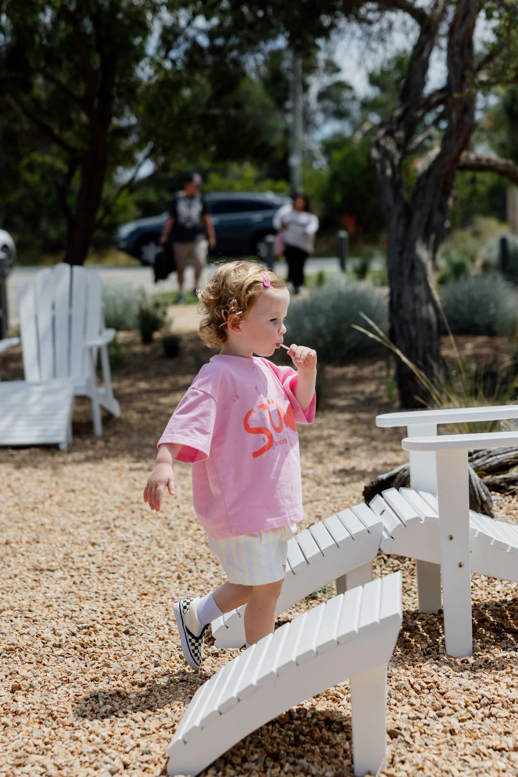 A curly-haired toddler strolls on a white wooden garden path, wearing an oversized fit of the SUNDAY SIBLINGS Il Sole Tee in Pink/Red made from 100% cotton, perfectly paired with light shorts. Adults and trees fill the background.