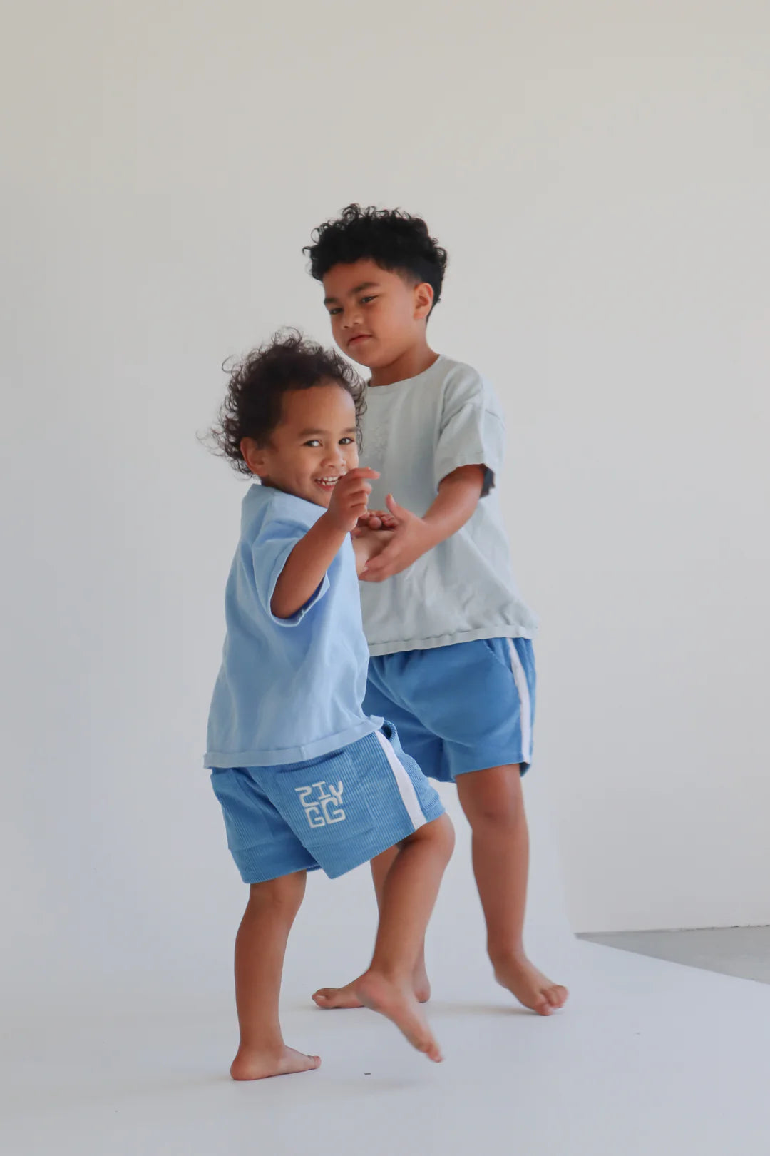 Two young children are joyfully playing together in a minimalist indoor setting with a white backdrop. They are both barefoot and wearing ZIGGY LOU's Cord Shorts River with an elastic waistband; one wears a light blue shirt while the other sports a Milk Stripe light gray shirt. Both children look happy and energetic.