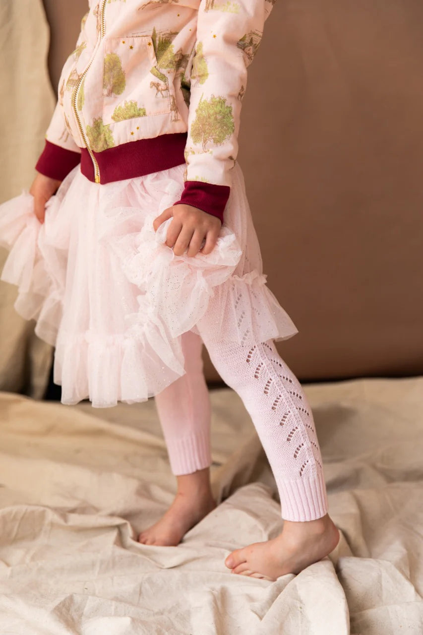 A young girl wearing the knitted leggings in lolly colour standing on cream drapes. She is wearing a zip up jacket with trees, horses, and old houses on it with a maroon trim around the waist and wrists.