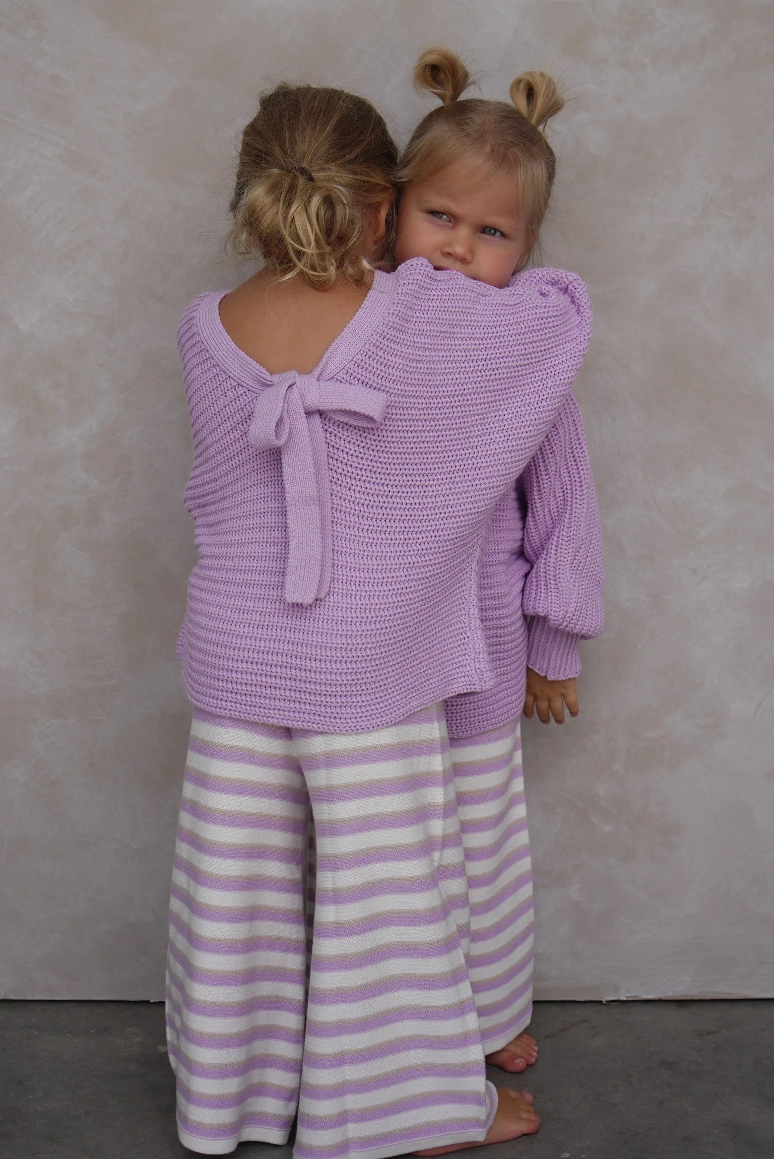 Two children wear matching EN. THE LABEL outfits: one child faces forward in the En. The Label ~ Stripe Knit Pants Lilac with flared bottoms, while the other shows off a lavender sweater with a back bow against a plain, light background.