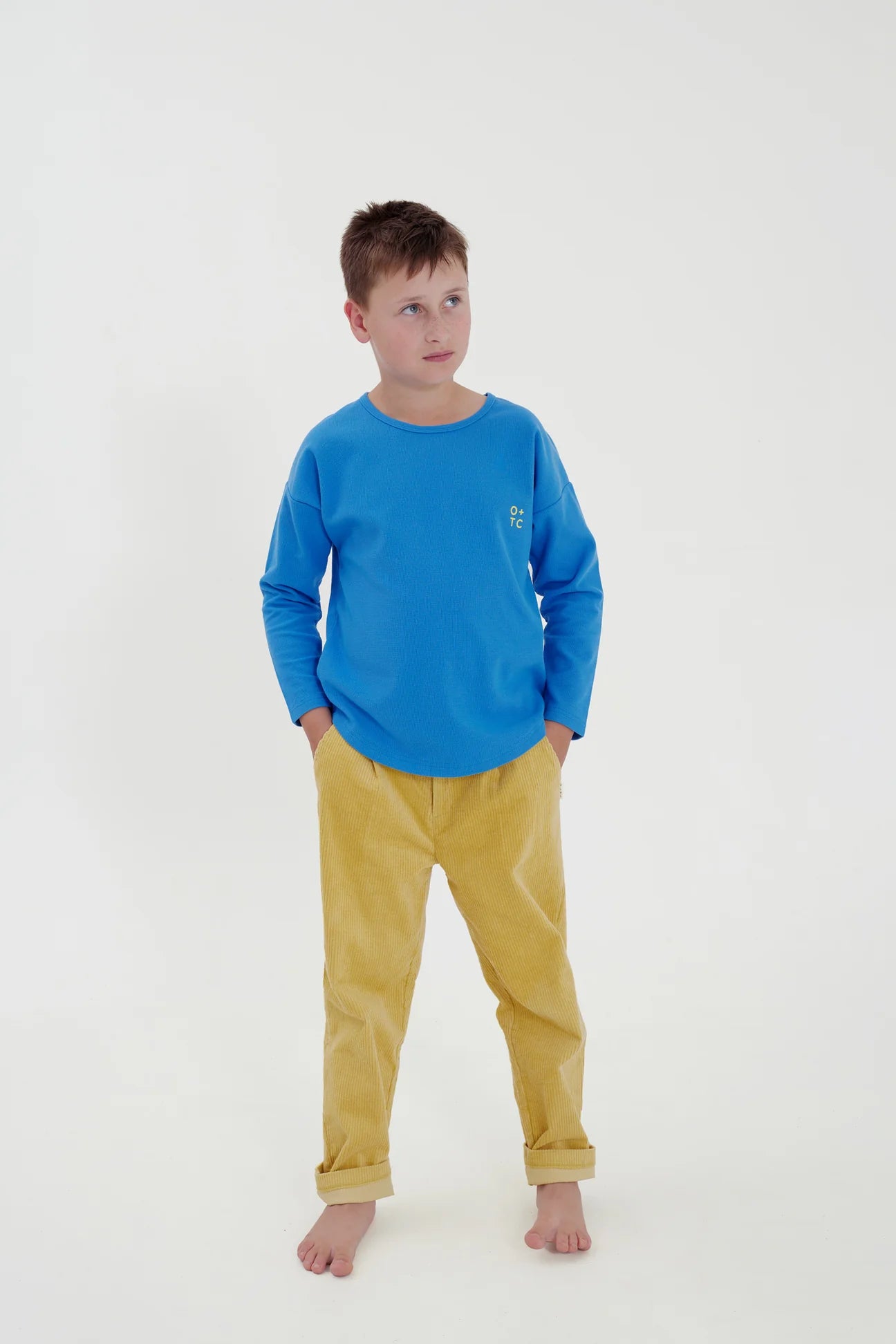 A young boy stands against a plain white background, wearing an O+TC Blue Relaxed Fit Long Sleeve Top by OLIVE + THE CAPTAIN and yellow pants made from a premium cotton polyester spandex blend. His hands are tucked into his pants pockets, and he is barefoot, looking slightly to the side with a relaxed expression.