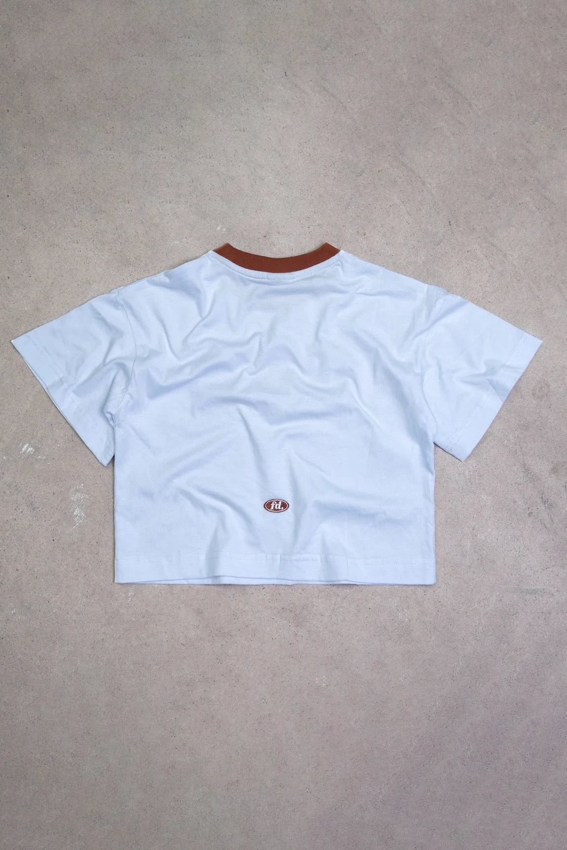 The PRE-ORDER Hibiscus Tee by FRANCO'S DAD is displayed flat on a concrete surface, highlighting its vintage 90's sports design. This light gray cropped T-shirt with a dark brown collar features a small oval-shaped logo near the lower back.