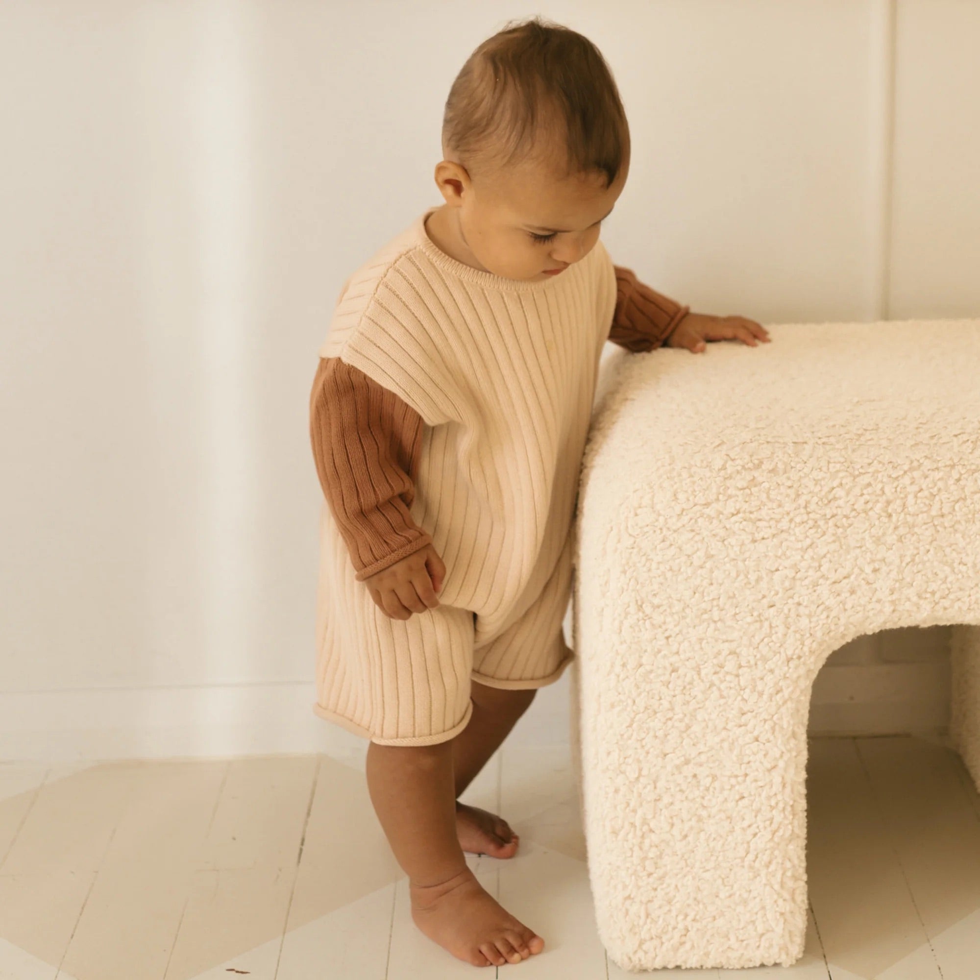 A toddler stands next to a soft, textured beige bench, wearing the Golden Knit Romper Caramel Sundae from GOLDEN CHILDREN. The child appears to be looking down, with one hand resting on the bench for support. The space has a warm, minimalistic feel with light-colored flooring and walls.