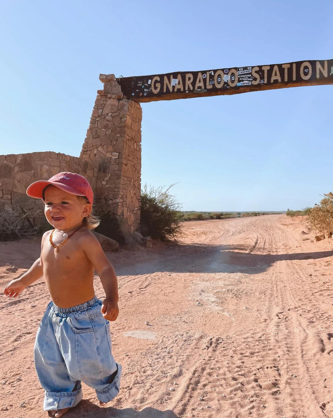 A young child in Ziggy Zaza - Suki Denim Jean Light Wash Denim by ZIGGY ZAZA, red cap, and beaded necklace walks barefoot under the "GNARALOO STATION" sign, with a stone arch and desert landscape in the background.
