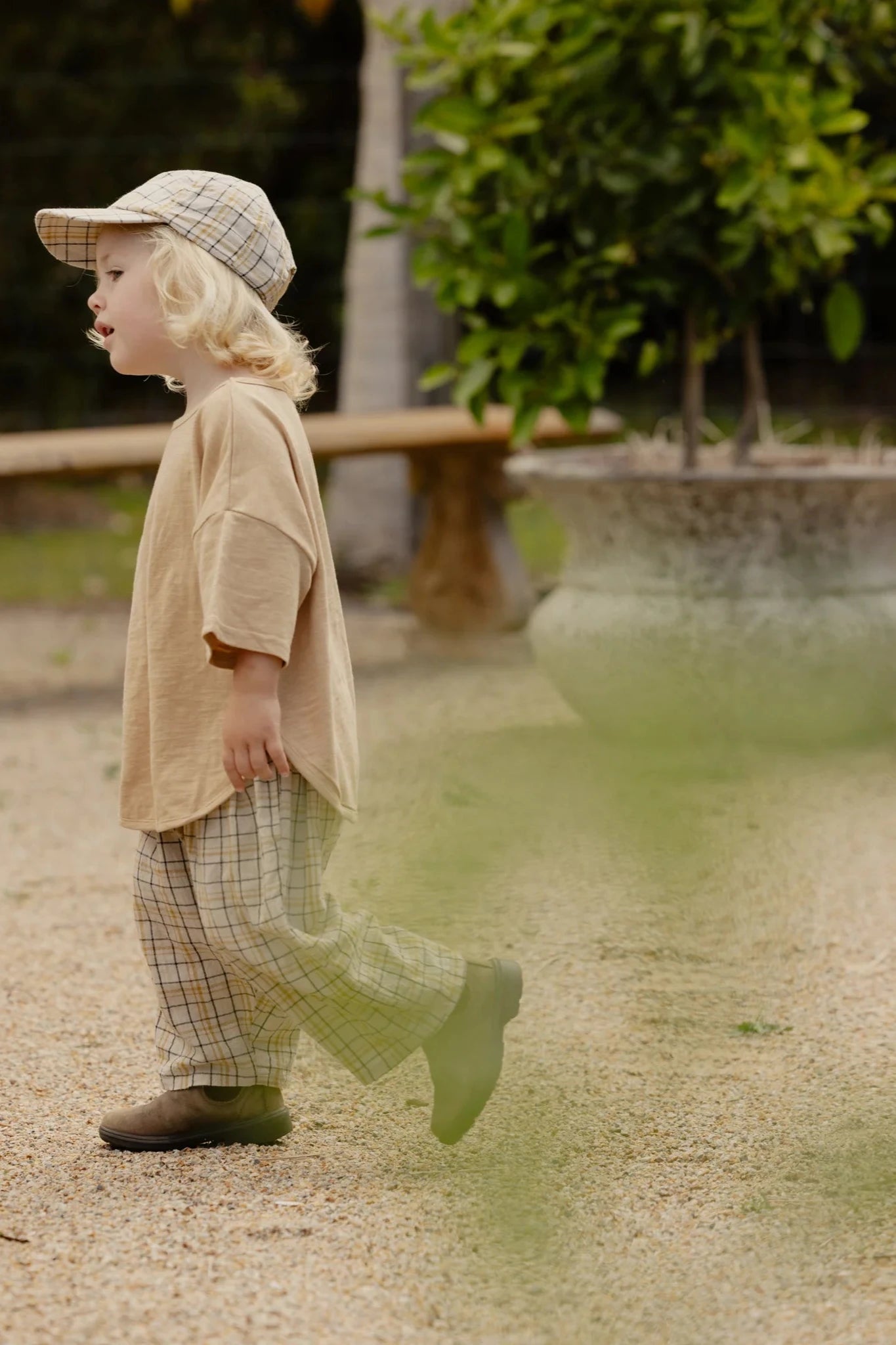 A small child with blond hair walks on a gravel path, dressed in a beige shirt and the VALENCIA BYRON BAY Rio Pant Picnic—practical plaid pants made of 100% cotton—with a matching plaid cap. A large planter with green foliage and a wooden bench are in the background, making the scene look like a park or garden.