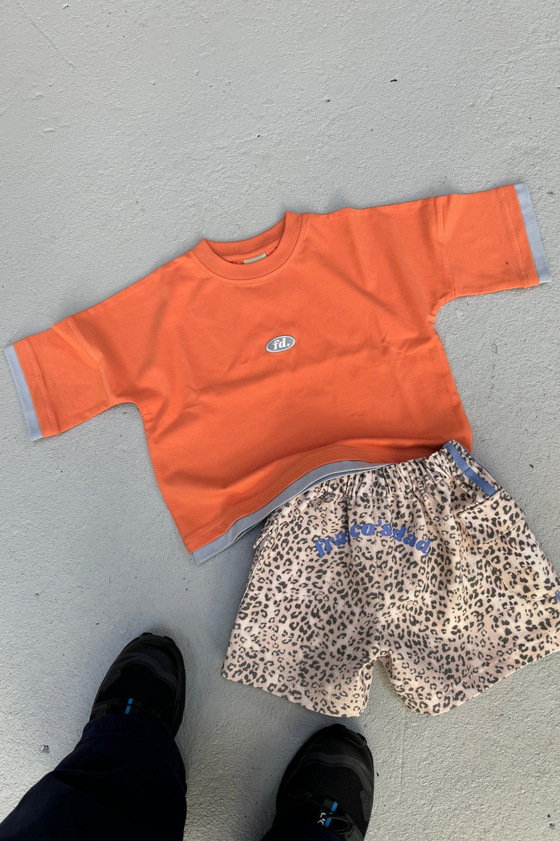 The PRE-ORDER Double Layer Tee Paw Paw from FRANCO'S DAD, featuring a subtle emblem on the chest, is paired with leopard print shorts accented with blue text. Black shoes peek out at the bottom, completing this oversized look.