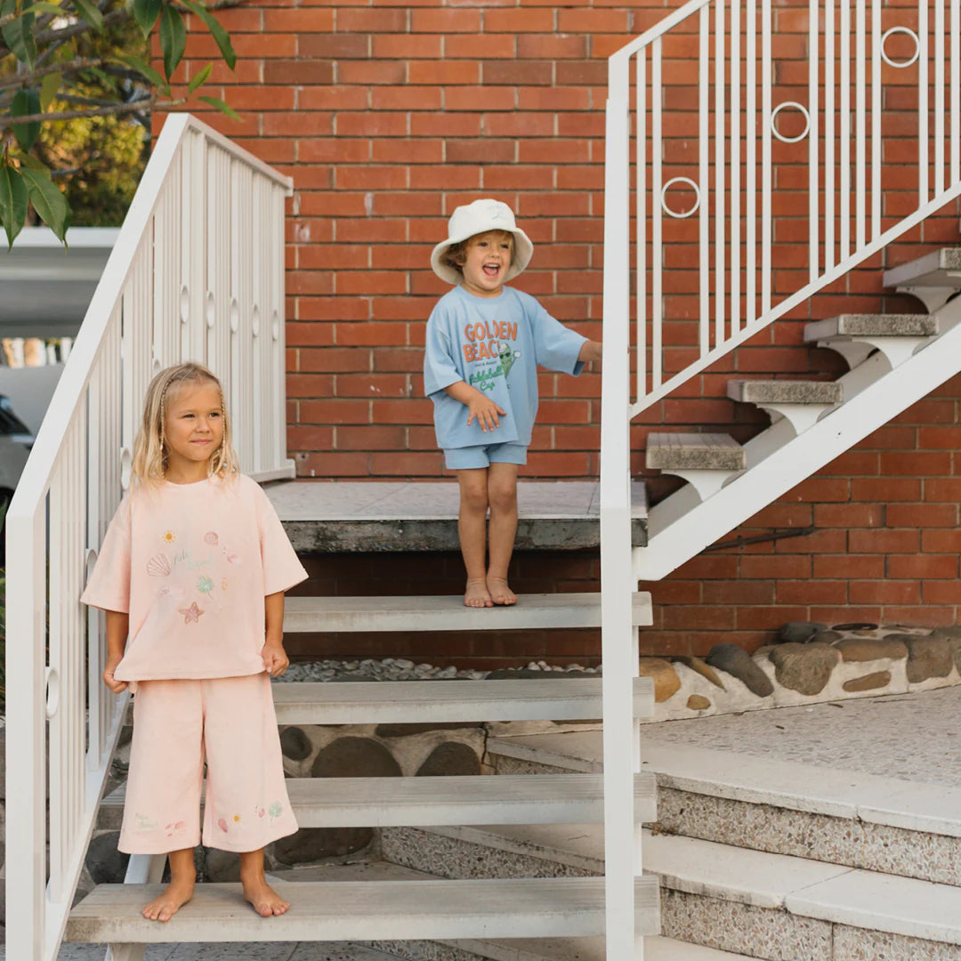 Two children stand on outdoor stairs in front of a brick wall. The girl, dressed in GOLDEN CHILDREN's Palm Beach Wide Leg Pants Terry Towel in Flamingo Pink, is on a lower step, while the boy in a blue shirt and white hat cheerfully stands above her. White railings border the stairs, capturing the essence of their playful moment.