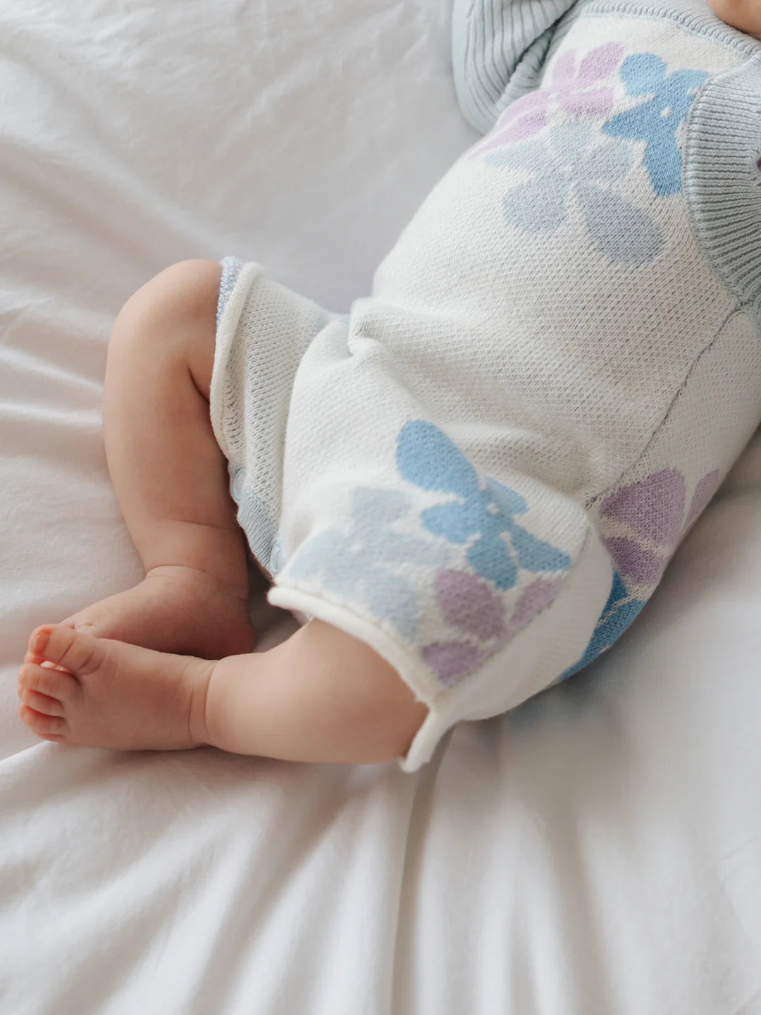 A baby wearing a light-colored ZIGGY LOU Playsuit Alaska with blue and purple floral patterns is lying on a white bed. The image focuses on the lower half of the baby's body, highlighting their legs and tiny feet.