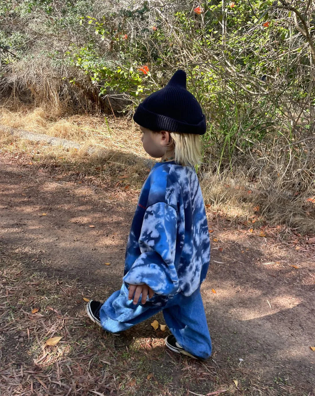 A young child in the ZIGGY ZAZA Ziggy Zaza - Toro Dyed Pullover Ocean, baggy jeans, and a black beanie walks along a dirt path with greenery and dry grass; the 100% cotton fleece adds comfort to their playful look.