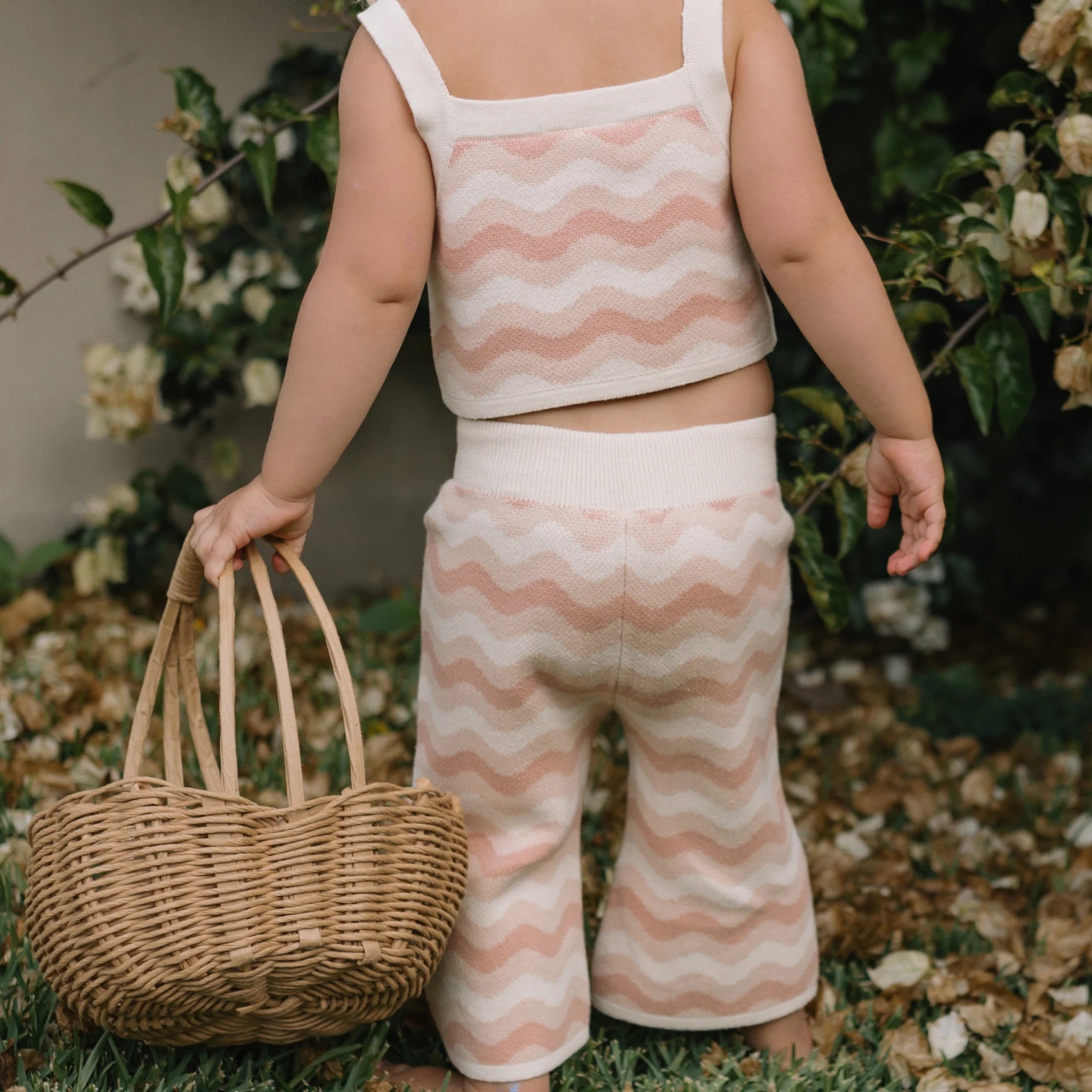 A toddler wearing the GOLDEN CHILDREN Seashore Knit Set in Strawberry Cream, featuring a sleeveless, relaxed-fit cotton design with a pink and white wavy pattern, holds a wicker basket while standing on grass next to a bush adorned with white flowers.