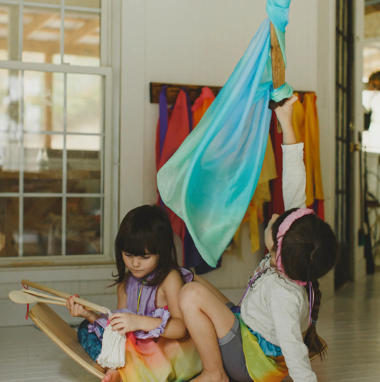 Two children are discovering their creativity indoors by a window. One is seated with a vibrant toy, while the other reaches up to the Enchanted Playsilk Sea from SARAH'S SILKS, with its beautiful blue and green hues. The room is adorned with vibrant cloths hanging in the background, encouraging unlimited imaginative play.