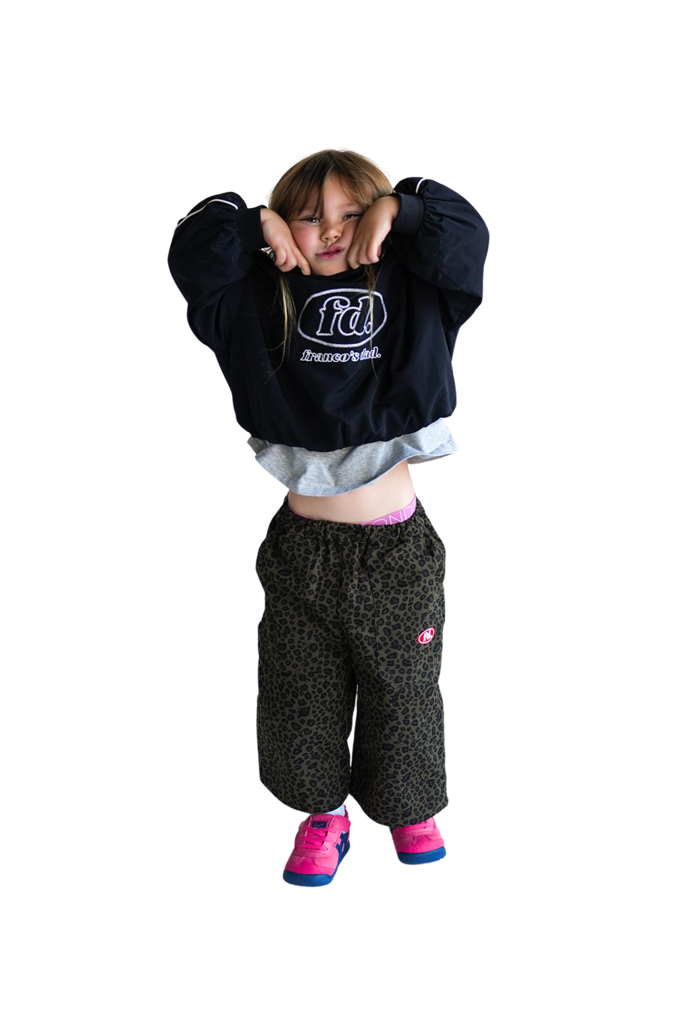 A young child playfully poses, pulling the hoodie drawstrings, wearing a black hoodie with white text, oversized PRE-ORDER Leopard Cargos from Franco's Dad, and pink shoes against a plain white background.