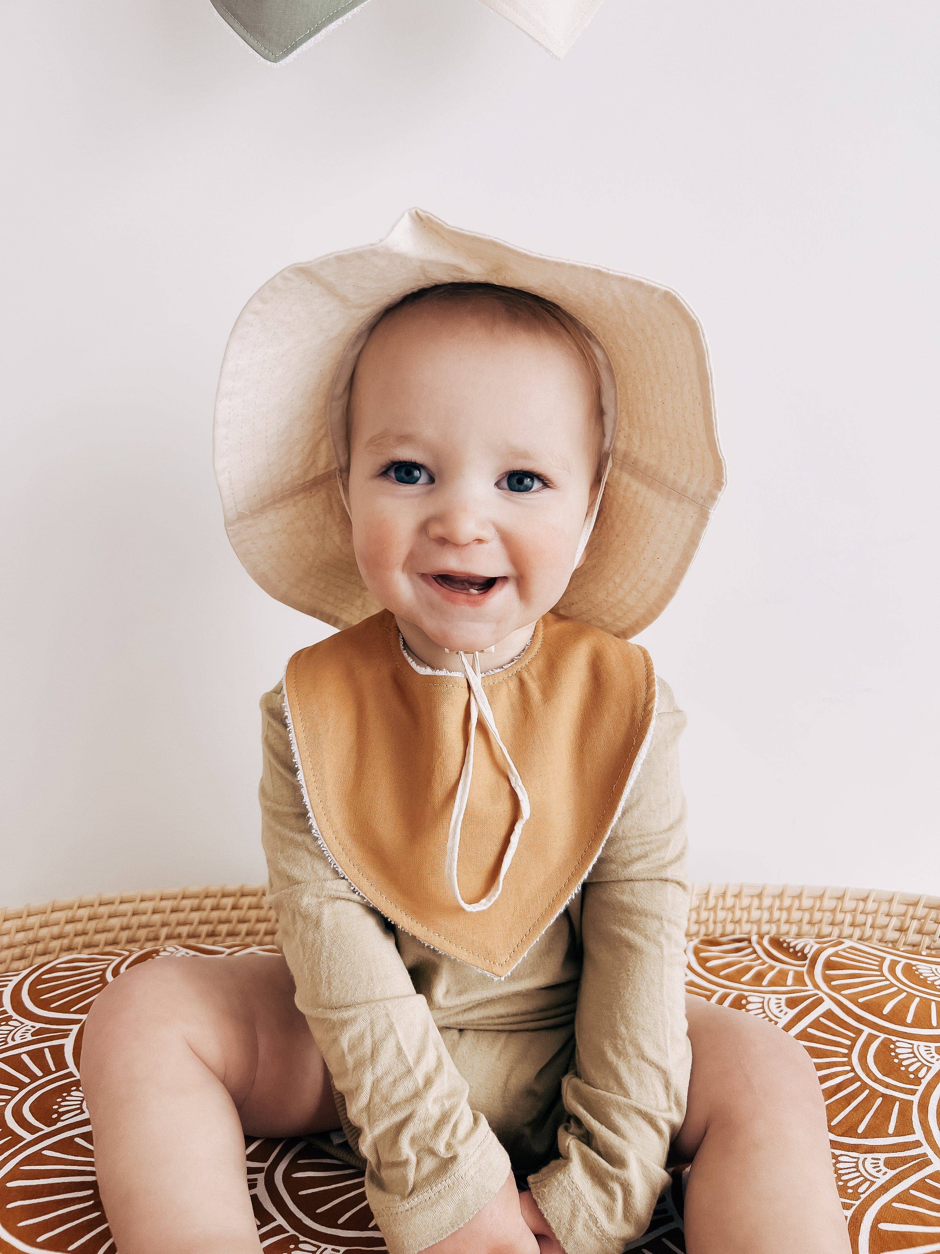 A smiling baby in a beige hat and Kiin Baby's Dribble Bib is seated on a patterned surface. The absorbent bamboo/cotton bib complements the baby's matching beige clothing against a light background.