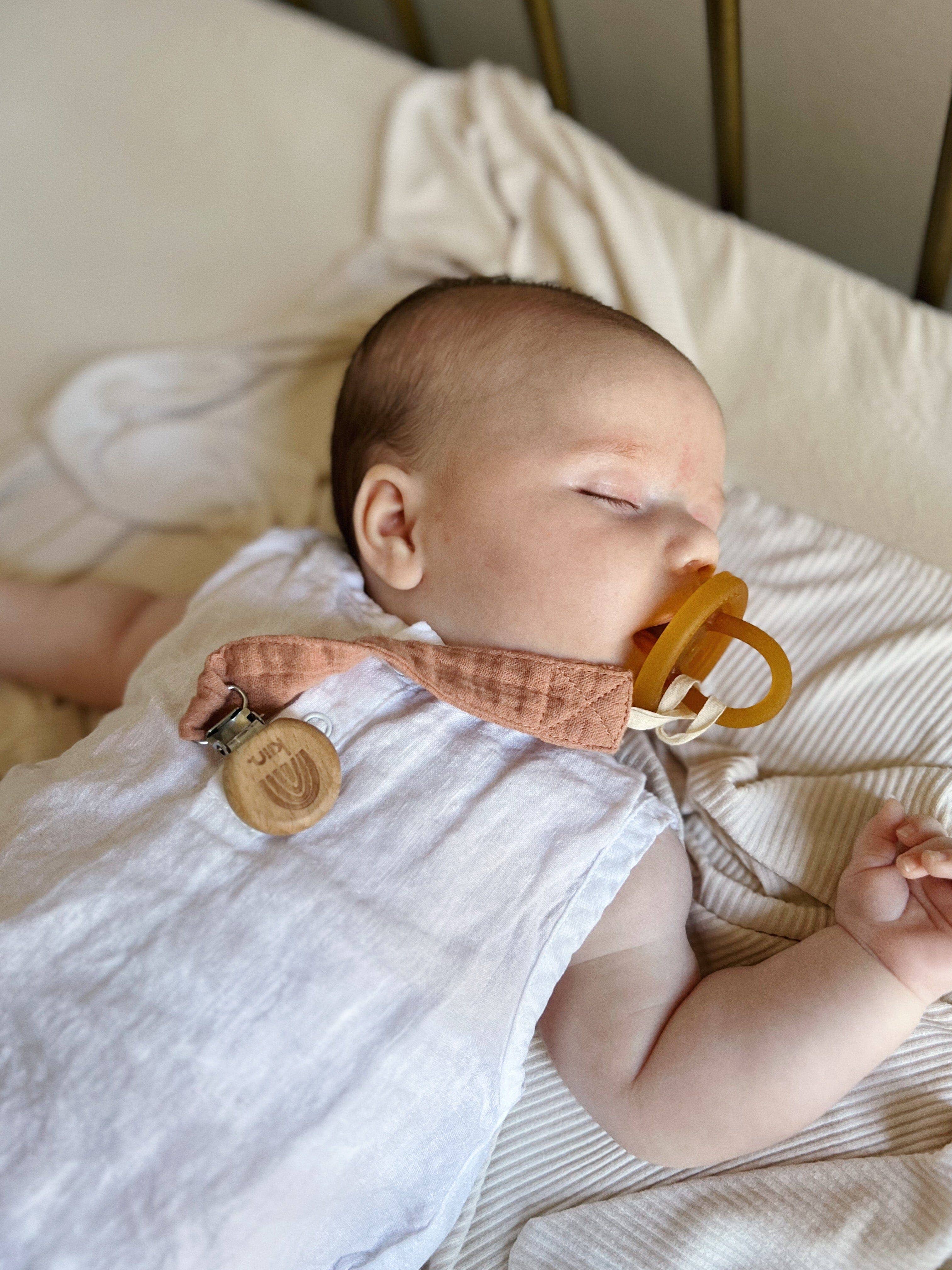 A baby in a white outfit peacefully sleeps with a KIIN BABY Cotton Dummy Clip Oat, ideal for gender-neutral nurseries. The subtly striped, light-colored bed linens create the perfect setting for this adorable baby shower gift.