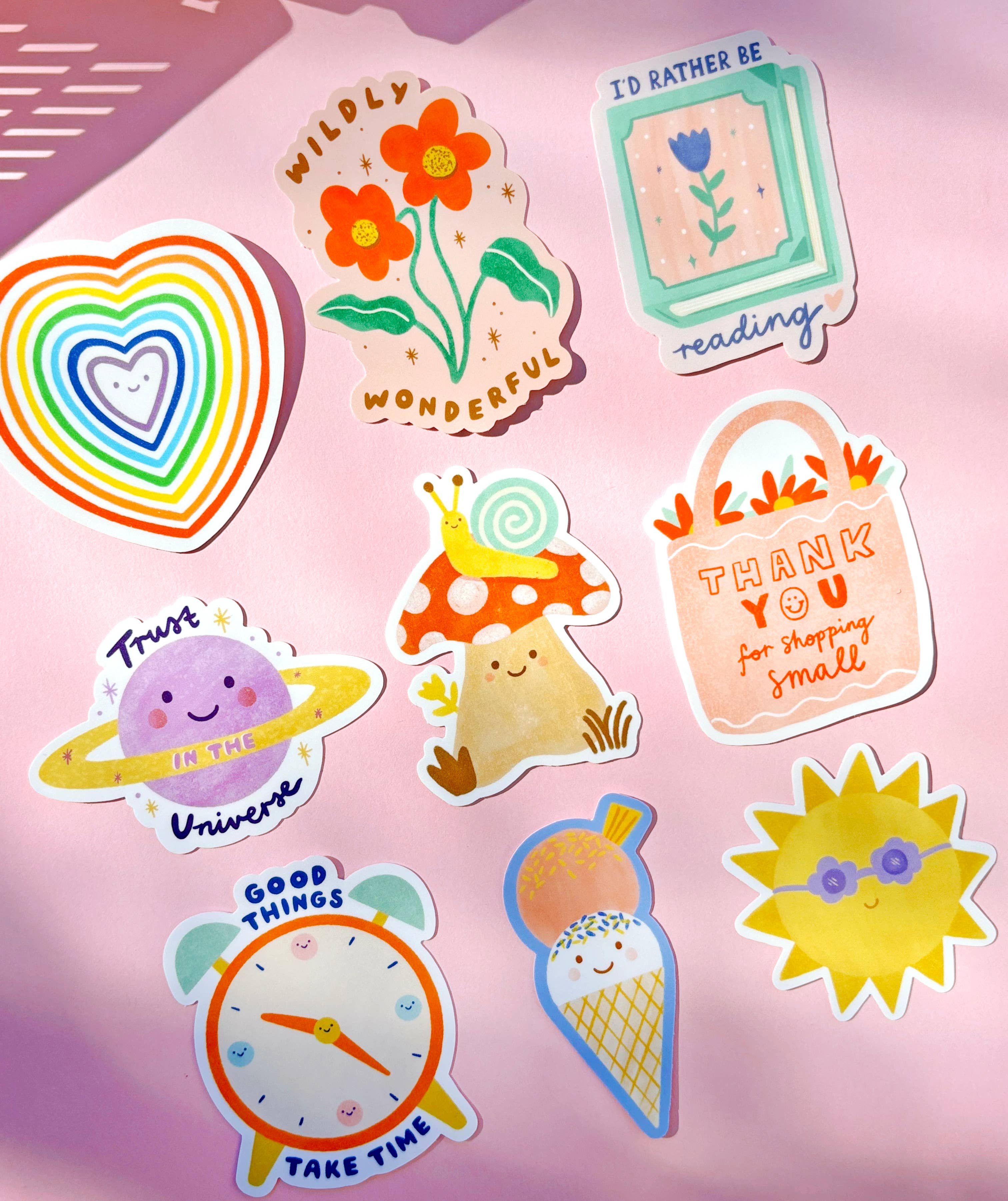 Introducing the Sunshine Sticker collection by LAUREN SISSONS STUDIO: A vibrant array of weatherproof vinyl stickers displayed on a pink surface, featuring designs like a rainbow heart, flowers, a cheerful sun with sunglasses, a whimsical mushroom, an ice cream cone, a clock, a book that says "I'd rather be reading," a thank you shopping bag, and a planet with text.