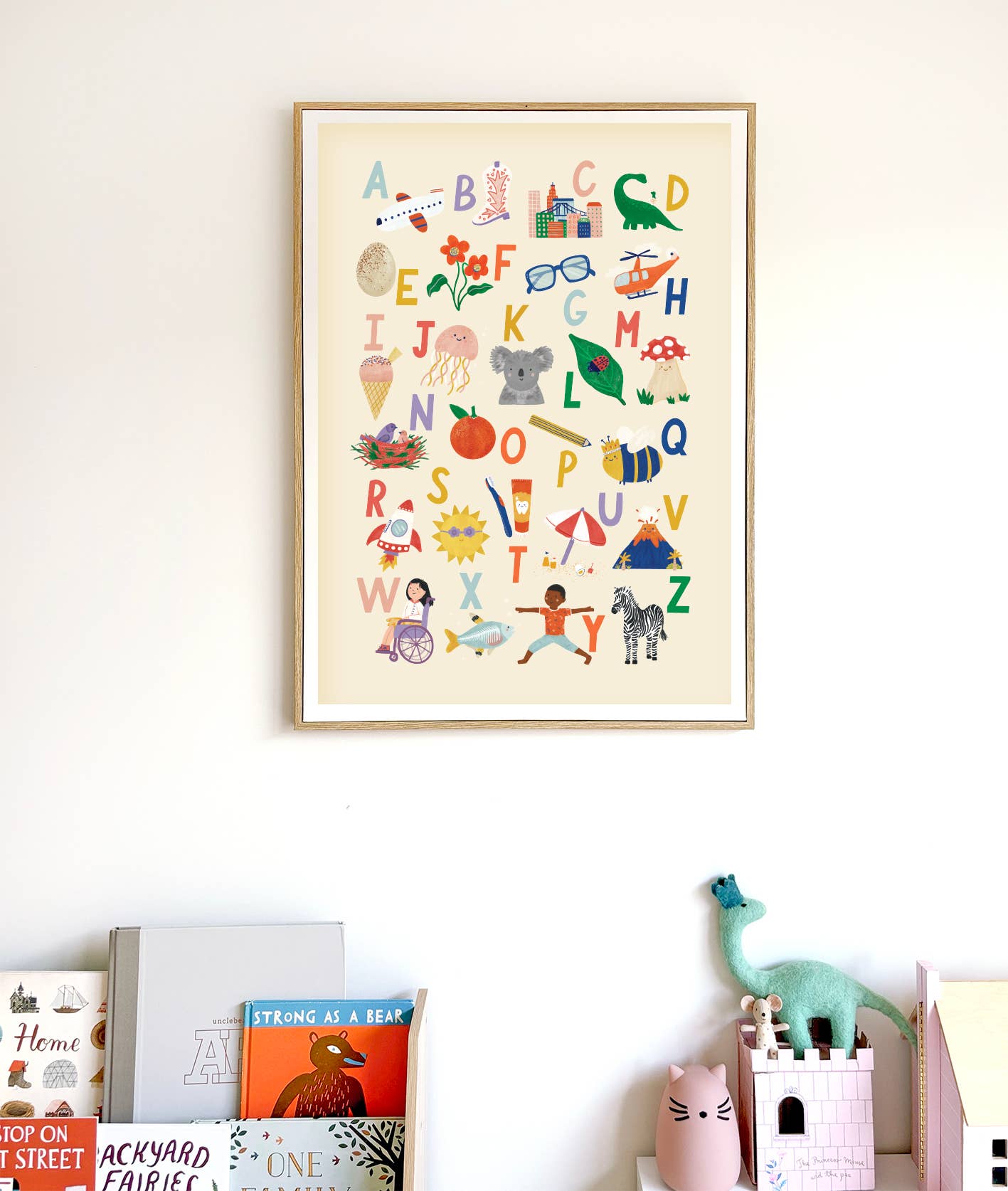 An Illustrated Alphabet Wall Art Print by LAUREN SISSONS STUDIO, showcasing playful illustrations for each letter, hangs on a white wall. Below it is a shelf adorned with various children's books, a green dinosaur toy, and a pink cat-shaped toy container. The nursery decor setup exudes a cozy and playful atmosphere.