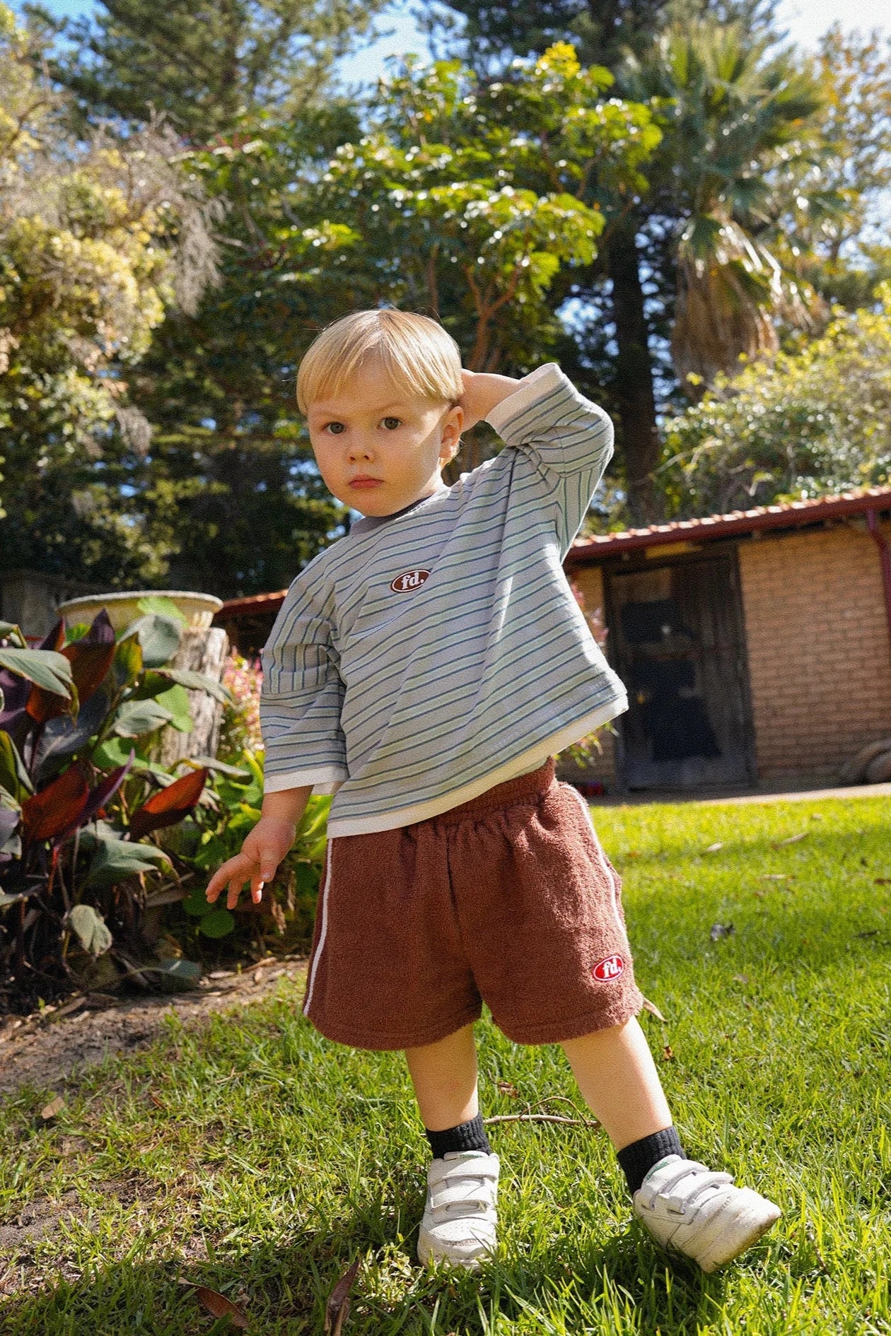 A small child with blond hair stands on the grass, dressed in a striped shirt and FRANCO'S DAD's PRE-ORDER Towel Shorts, which have a relaxed fit, paired with white shoes. They're touching their head amidst plants with a building in the background beneath a clear, sunny sky—capturing an ideal summer staple moment.