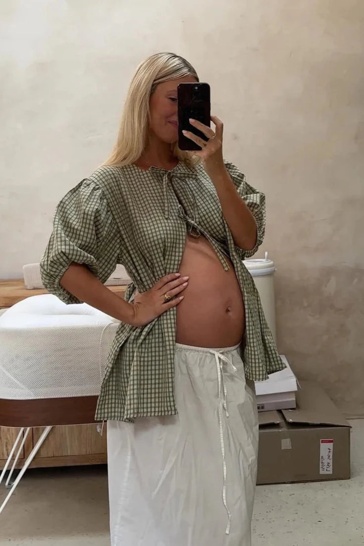 A woman with long blonde hair takes a mirror selfie, proudly showing off her pregnant belly. She's wearing the Mama Cami Blouse in olive gingham from LITTLE THE LABEL, paired with a white skirt. The room around her maintains a neutral tone background, including a bed and a cardboard box in sight.