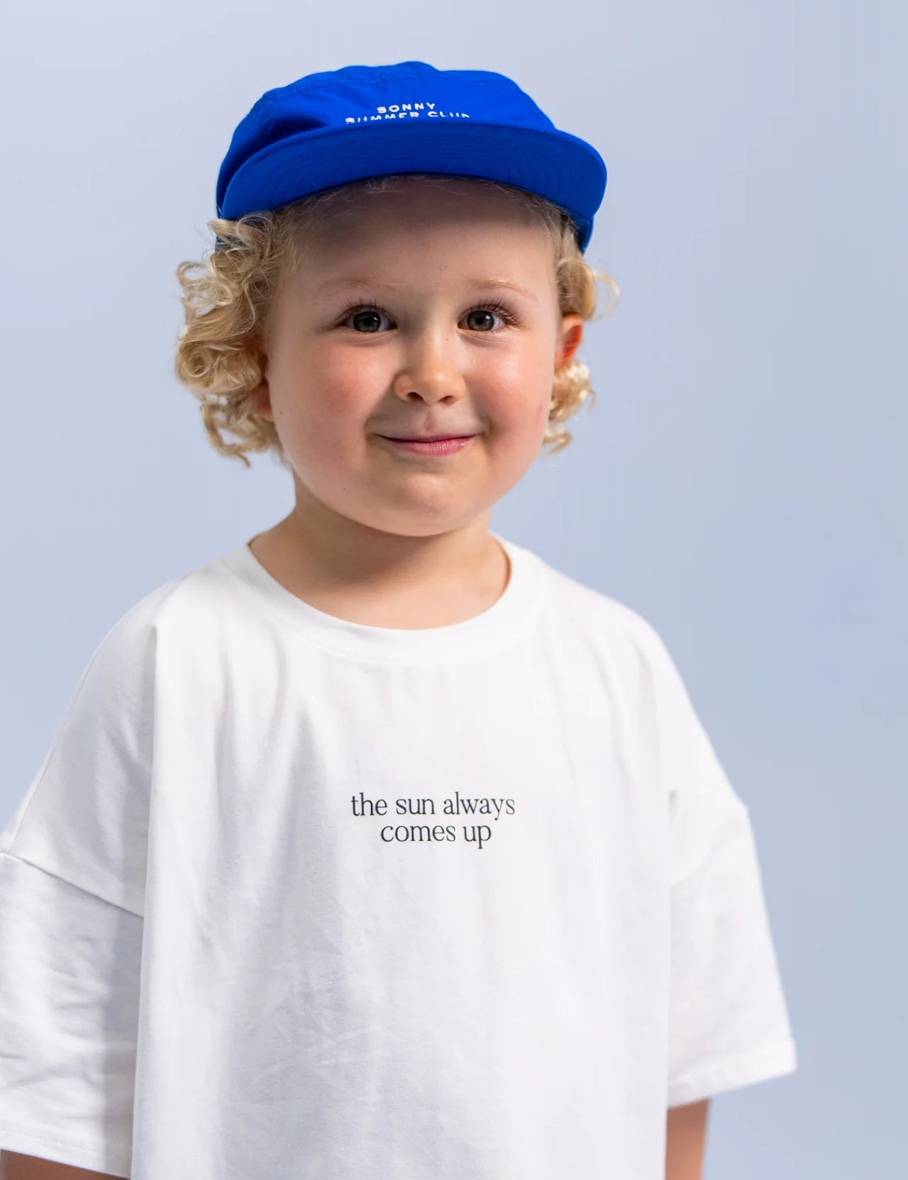 A young child with curly blonde hair beams while wearing the SONNY LABEL Summer Club Cap in Electric, which features an adjustable velcro strap, and a white T-shirt that says "the sun always comes up." The backdrop is a gentle blue.