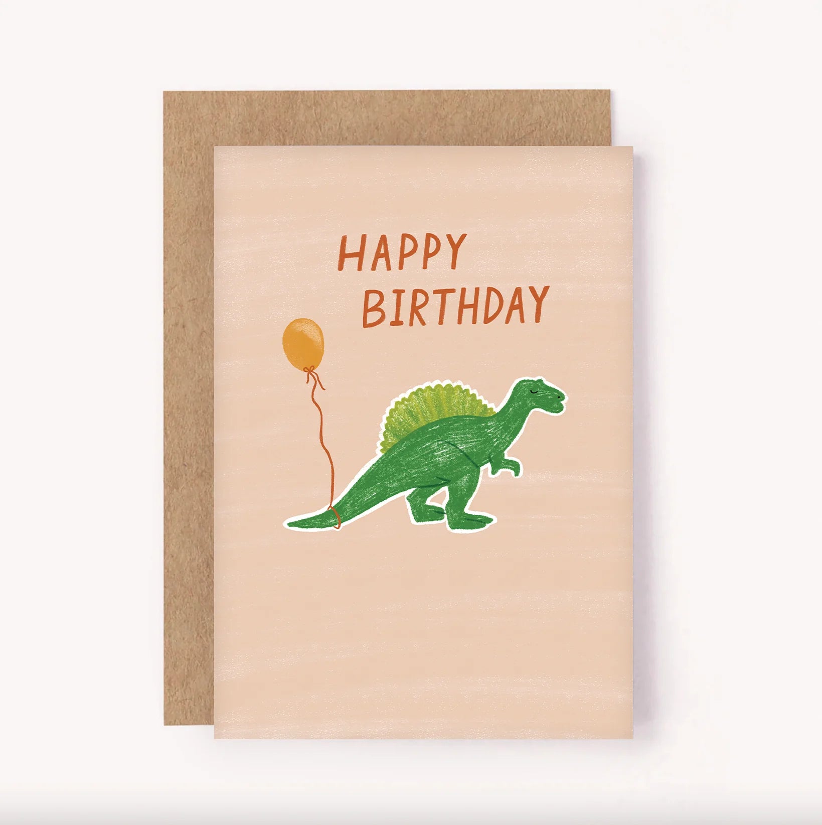 Introducing the Birthday Greeting Card “Dinosaur Balloon” by LAUREN SISSONS STUDIO. This charming card has a light beige background showcasing a playful green cartoon dinosaur holding an orange balloon. The text "HAPPY BIRTHDAY" is prominently displayed in orange letters at the top. The delightful illustration is layered on a slightly larger plain brown card, made of recycled paper, adding an eco-friendly touch to your celebration.