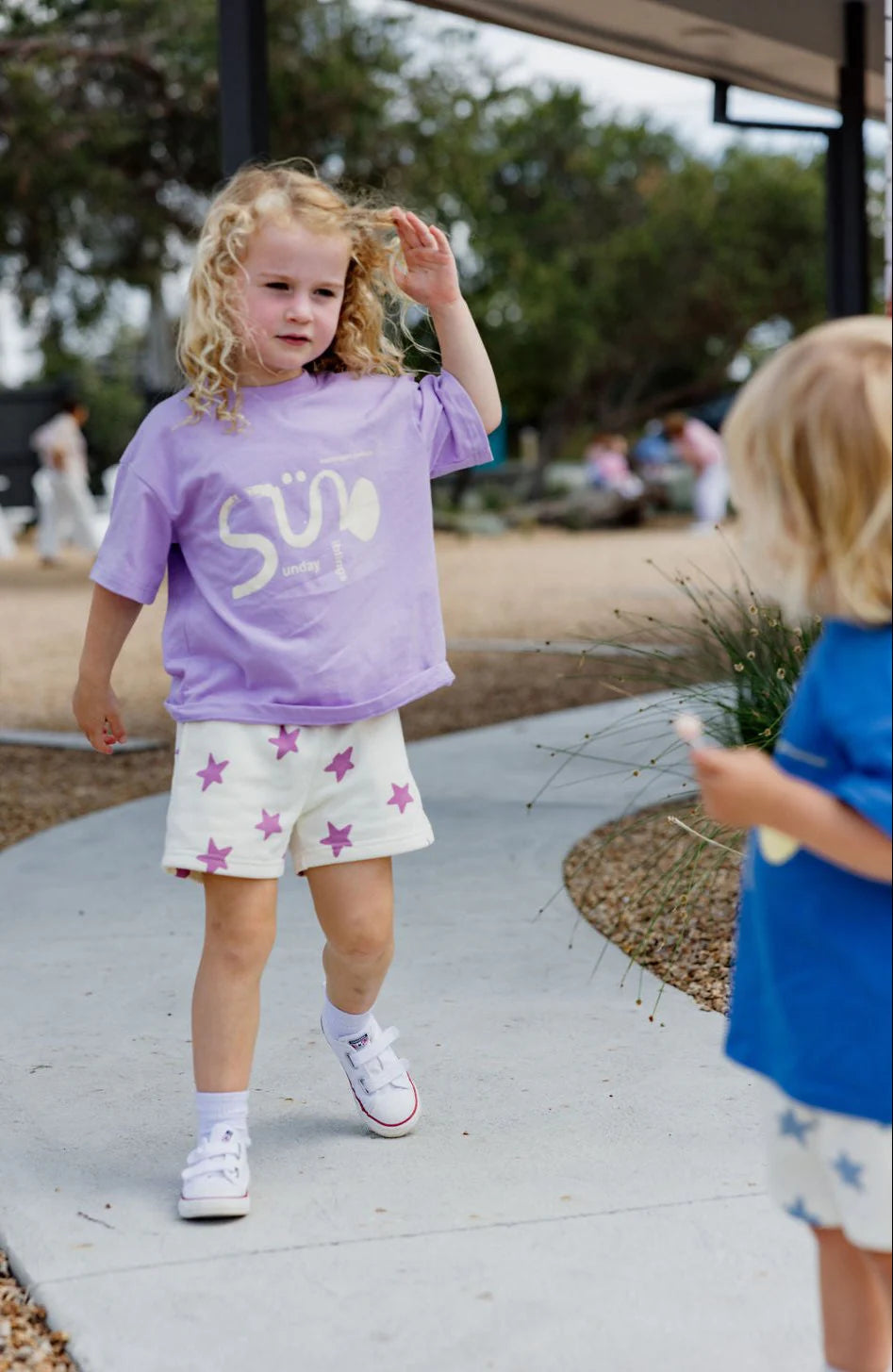 A curly-haired child in a purple shirt and SUNDAY SIBLINGS' Sunday Siblings ~ Kiddo Shorts Purple walks a curved path. Nearby, another child in a blue shirt wears similar French terry cotton shorts. The serene backdrop includes trees and people sitting.