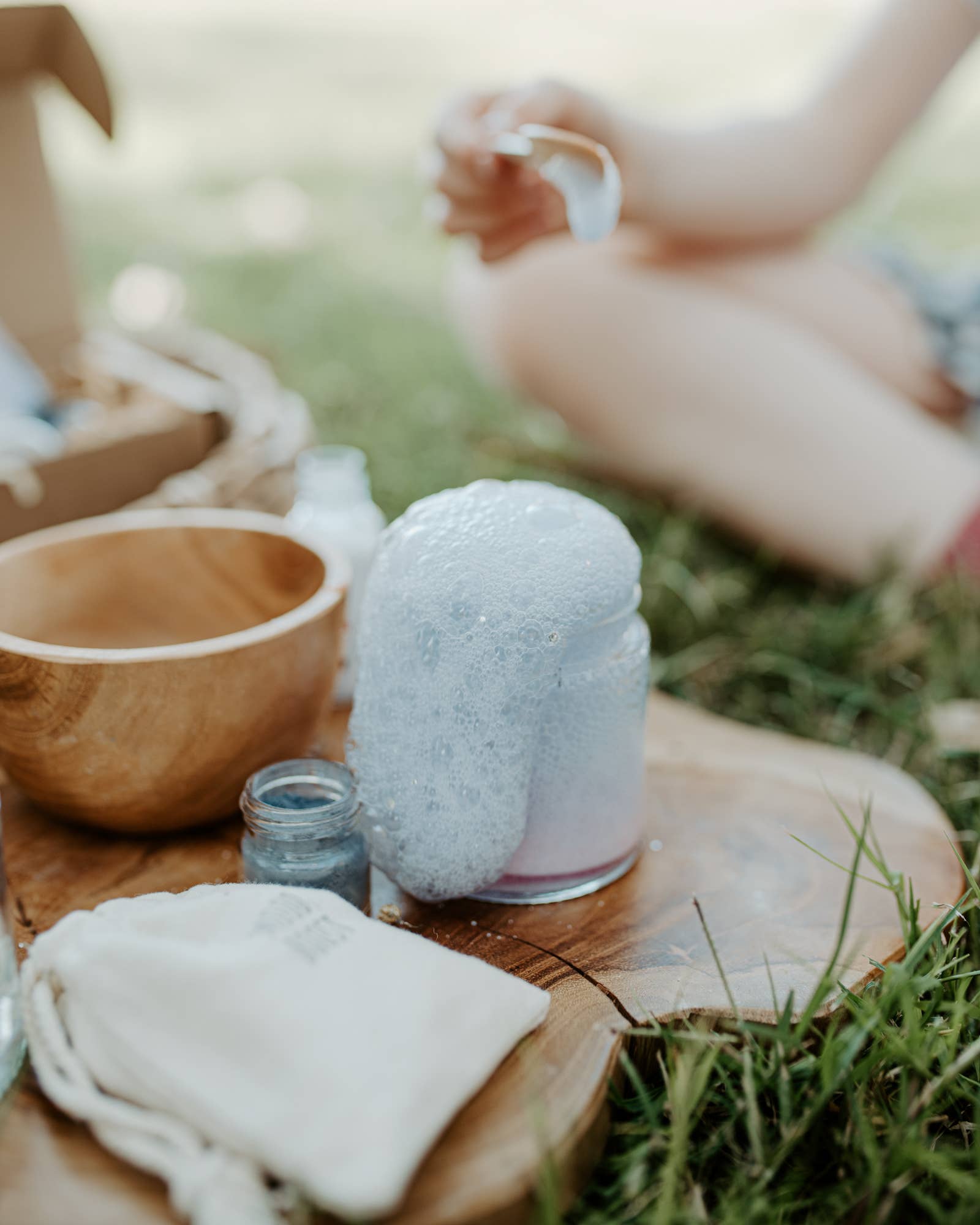 A close-up of a frothy science experiment in a jar outdoors, with bubbles overflowing onto a wooden surface. Nearby are small bowls, a jar of blue powder, and a cloth pouch from the MINI Colour Mood Potion Kit (Colour Changing Kit) by THE LITTLE POTION CO. A person is partially visible in the background, sitting on the grass.