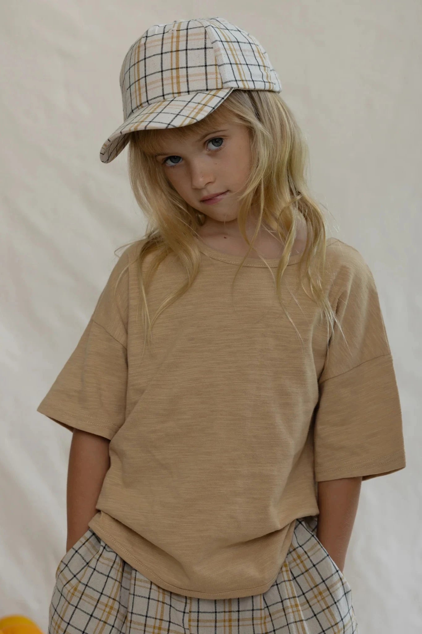 A young child with long blond hair wears a beige T-shirt and the Rio Pant Picnic from VALENCIA BYRON BAY, complemented by a matching plaid cap. The child stands against a plain light-colored background, looking directly at the camera with a slight tilt of the head, showcasing durable and practical unisex kids' clothing.