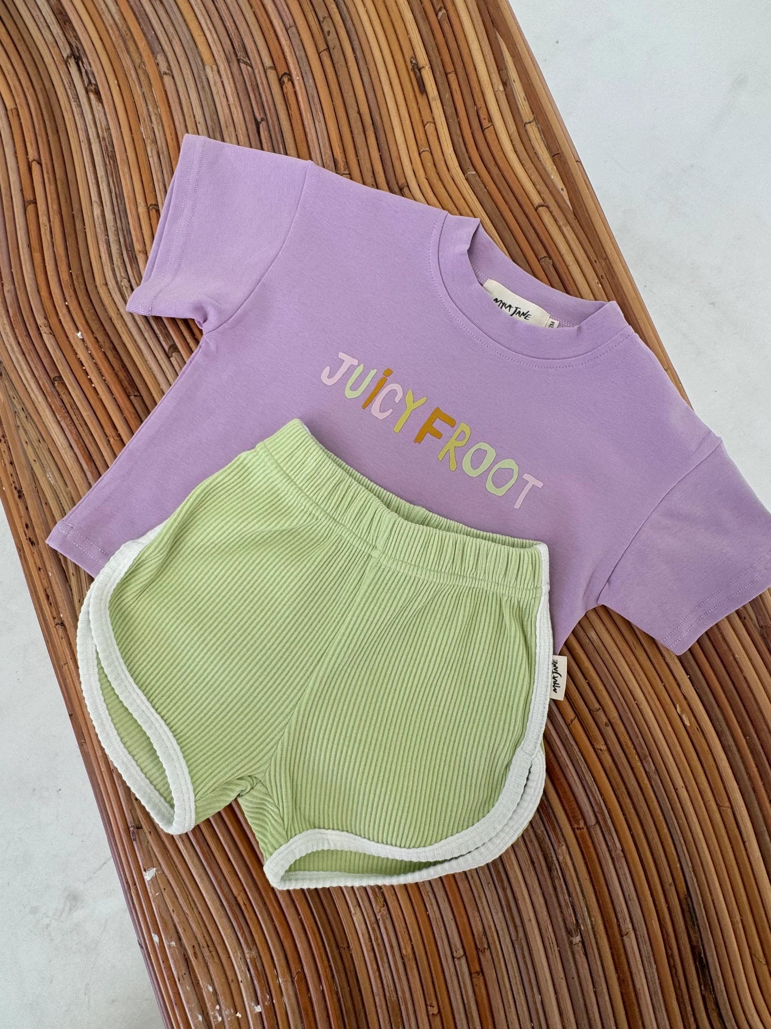 Displayed on a wooden surface is an adorable children's ensemble featuring the "Juicy Froot T-Shirt Grape" by MYLA JANE. This durable cotton purple T-shirt with bold "JUICYFRUIT" lettering perfectly complements the light green ribbed shorts, accented with white trim. It's a delightful choice for everyday play and comfort!