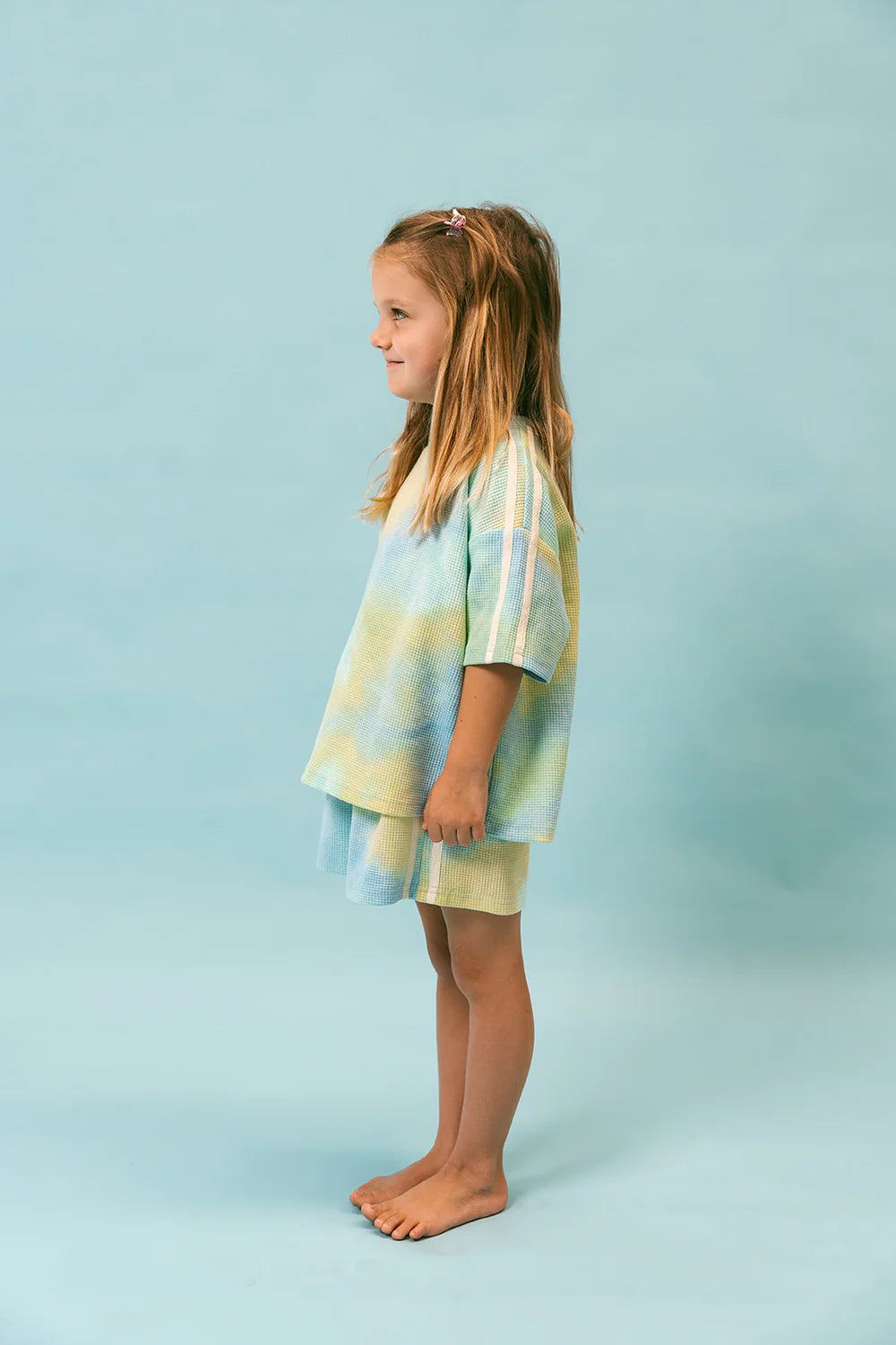 A young girl stands barefoot against a light blue background, dressed in an oversized "Waffle Set - Arctic" from LENN LABEL. The pastel-colored outfit features ribbed cotton fabric with hints of green, yellow, and blue. Her long, light brown hair is styled with a clip on one side, and she smiles gently.