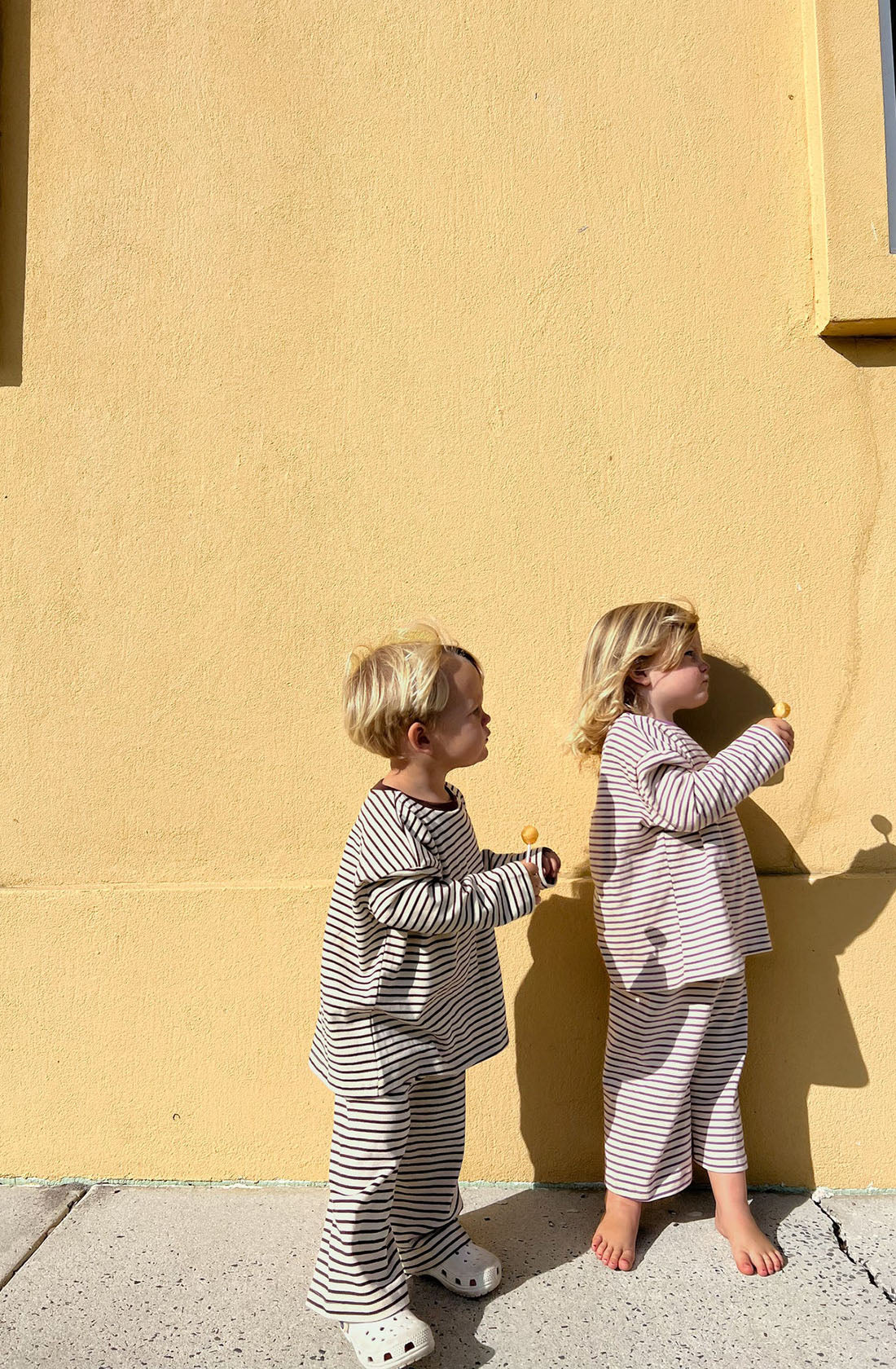 Two young children with blonde hair stand against a yellow wall, both holding ice cream cones. They are wearing matching Every Day Set Choc Chip outfits from LENN LABEL, made of 100% knit cotton. One child wears the brown and white striped outfit with white crocs, while the other wears the pink and white striped outfit and is barefoot.