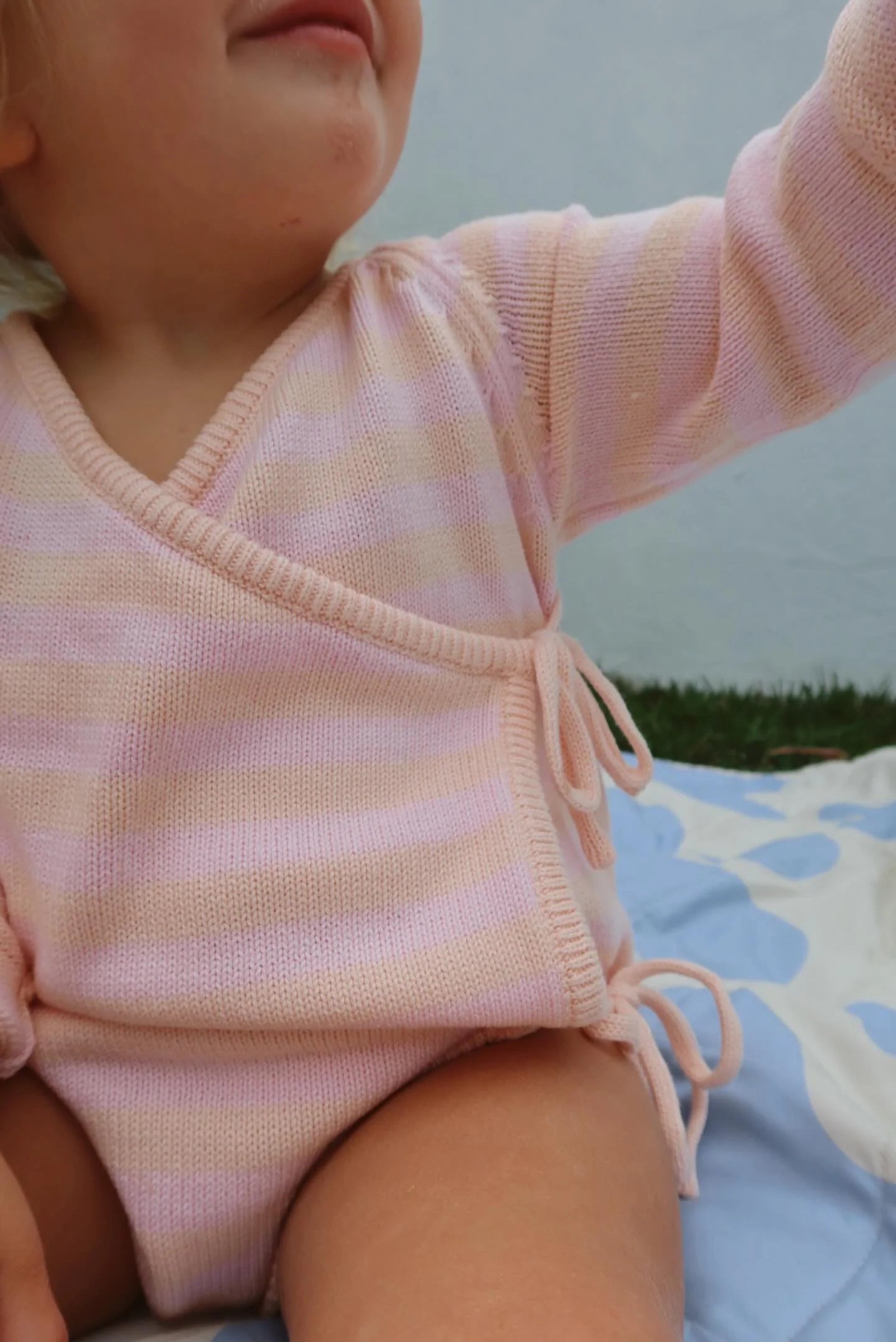 A close-up image of a baby wearing the MILKY DESIGNS Essential Wrap Romper in Pink/Peach, made from 100% cotton. The baby is sitting on a blanket with a light blue and white polka dot pattern, radiating comfort. The baby's face is partially visible, showing a smile, while the background is out of focus.