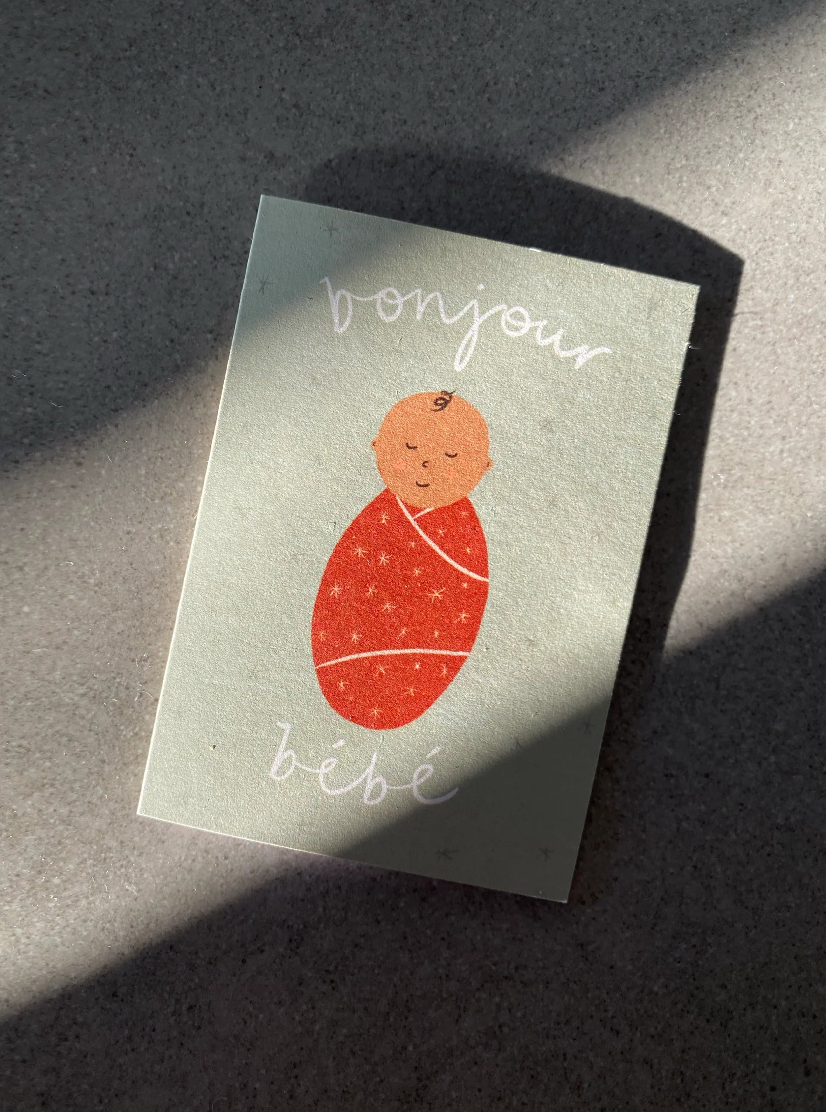 The Bonjour Bebe Mini Card - New Baby from LAUREN SISSONS STUDIO lies on a surface with sunlight casting shadows. The card features an illustration of a swaddled baby in an orange wrap adorned with tiny stars. The text "bonjour" appears above the illustration, and "bébé" is written below in a handwritten style.