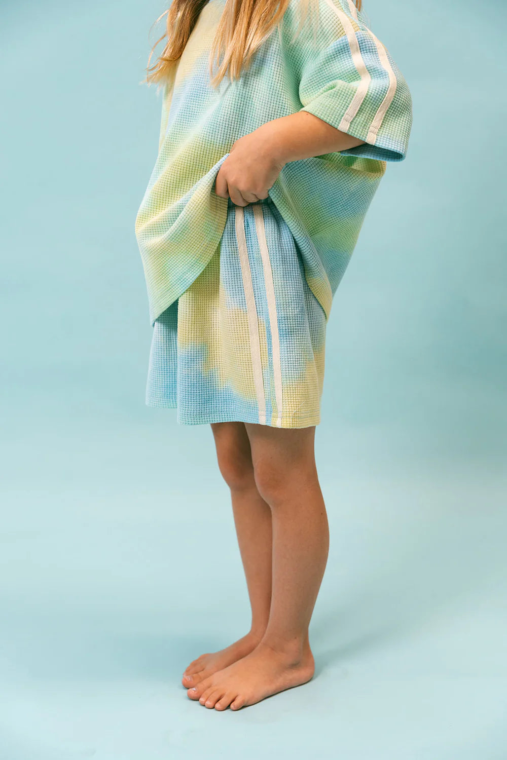 A young child stands barefoot against a blue background, wearing LENN LABEL's oversized "Waffle Set - Arctic," a pastel-colored outfit featuring stripes. The set includes a shirt and shorts patterned in light blue, green, and yellow hues and is crafted from comfortable ribbed cotton. The child holds one side of the shirt.