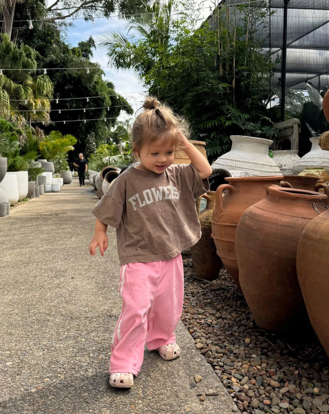 A young child with a topknot strolls along a path adorned with large clay pots and greenery. Wearing a relaxed boxy fit "Ziggy Zaza ~ Flowers Tee Sparrow" from ZIGGY ZAZA, pink pants, and bear-themed slippers, the scene features someone walking in the background.