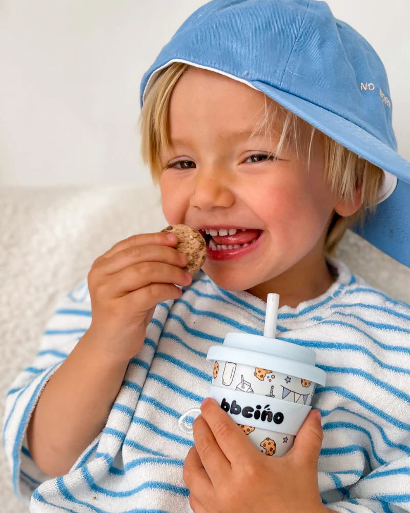 A smiling child in a striped shirt sports a blue cap while holding a cookie in one hand and BBCINO's Reusable Babycino Cup 120ml Tough Cookie with a reusable silicone straw in the other. The background features a soft, neutral shade.