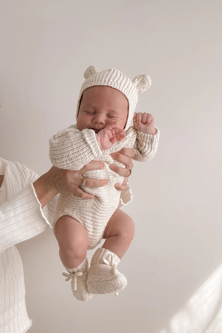 A person is holding up a baby dressed in the adorable cream-colored Heirloom Romper Honey from the ZIGGY LOU collection. The baby is wearing a knitted hat with bear ears, a matching onesie, and booties—a perfect ensemble for showcasing stylish baby clothing. The baby's face is scrunched up as they seem to be yawning or stretching. The background features a plain light color.