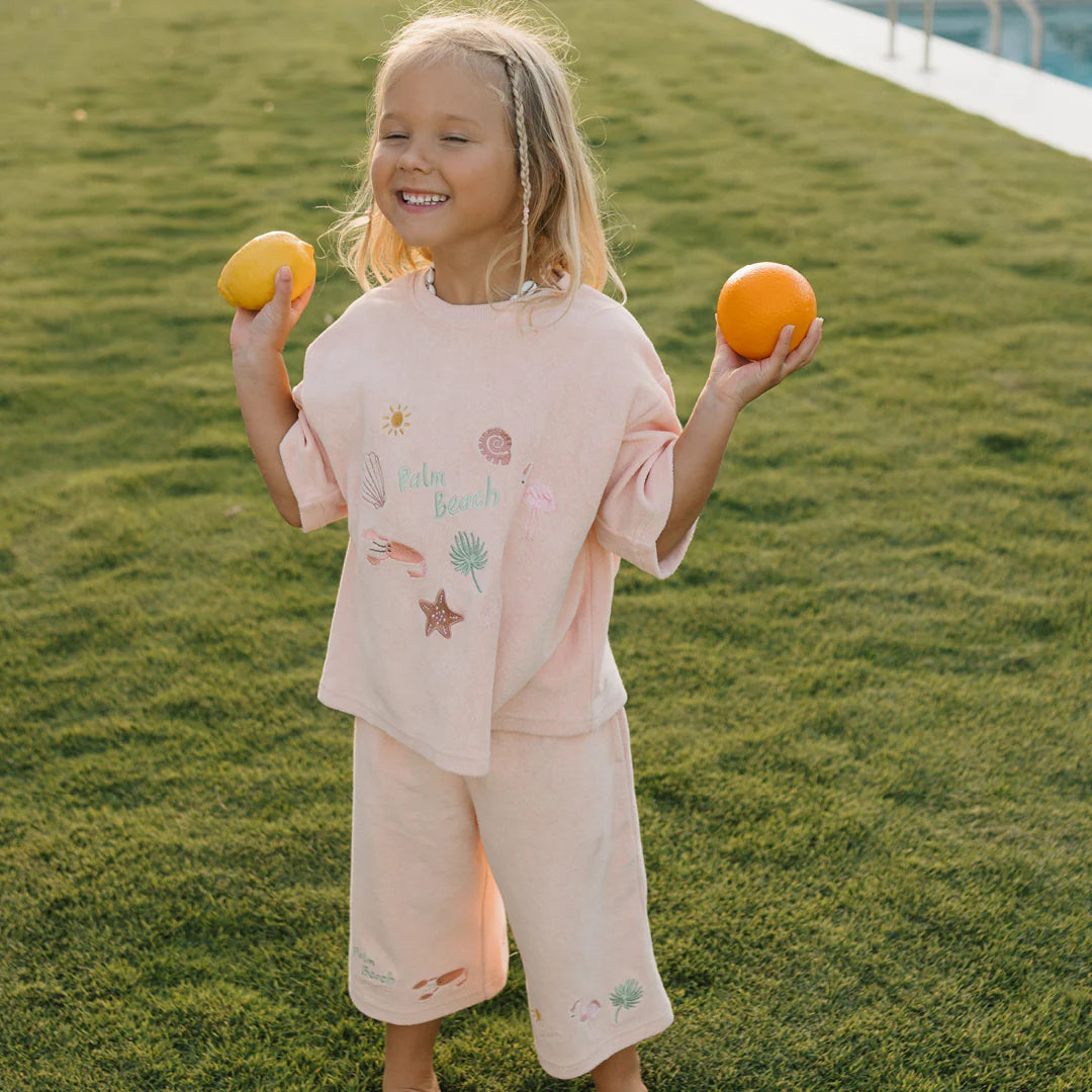 A young girl with a joyful expression, sporting blonde hair and dressed in a GOLDEN CHILDREN Palm Beach Mid Sleeve Tee Terry Towel in Flamingo Pink adorned with beach-themed embroidery, stands on grass holding an orange in one hand and a lemon in the other. A pool can be seen in the background.