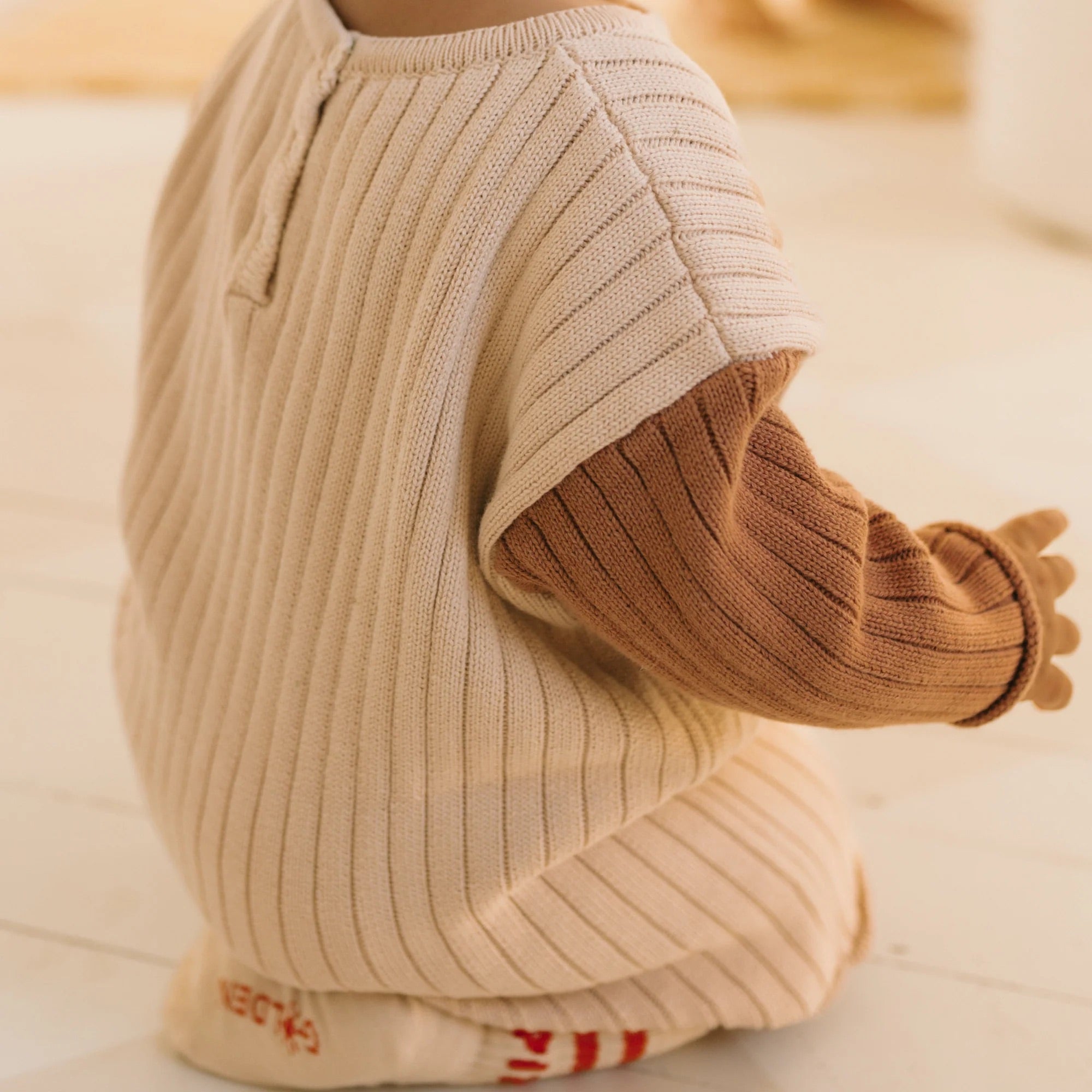 A young child, with their back to the camera, sits on the floor dressed in GOLDEN CHILDREN's Golden Knit Romper Caramel Sundae. They wear a ribbed, cream-colored short-sleeve sweater over a long-sleeve brown cotton knit rib sweater, complemented by cream socks featuring red stripes on the bottom.