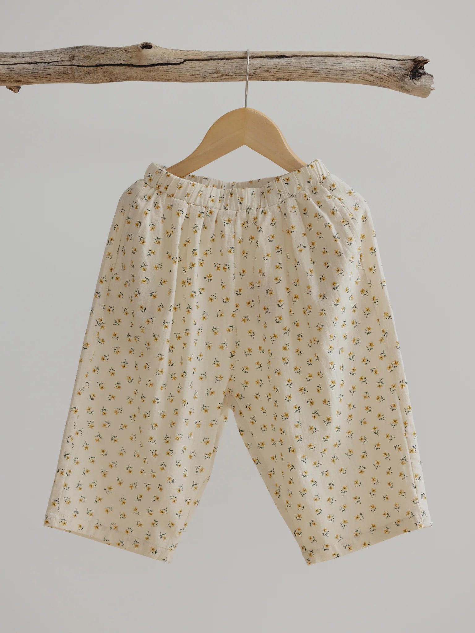 A pair of Rio Pant Marigold from VALENCIA BYRON BAY, light cream-colored and loose-fitting with a subtle floral pattern and hints of marigold, hangs on a wooden hanger suspended from a natural, rough-hewn wooden branch against a plain white background. Crafted from 100% cotton for comfort and durability, these versatile pants are perfect for any occasion.