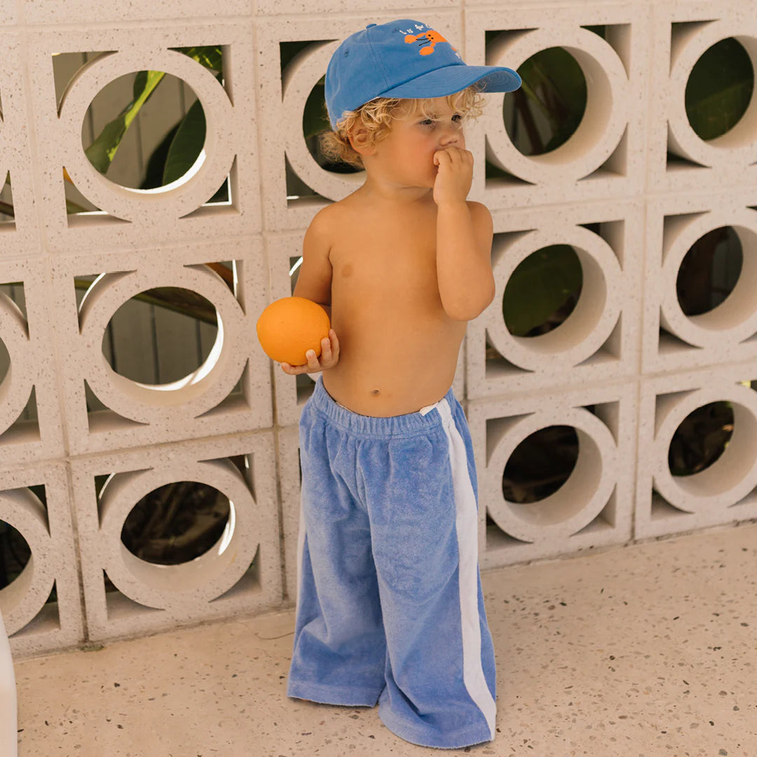 A toddler wearing a blue baseball cap and relaxed Pool Blue pants from Golden Children's Ciao Tutti collection holds an orange against a decorative white wall. Dressed comfortably in terry towel cotton, the child gazes to the side with a hand near their mouth.