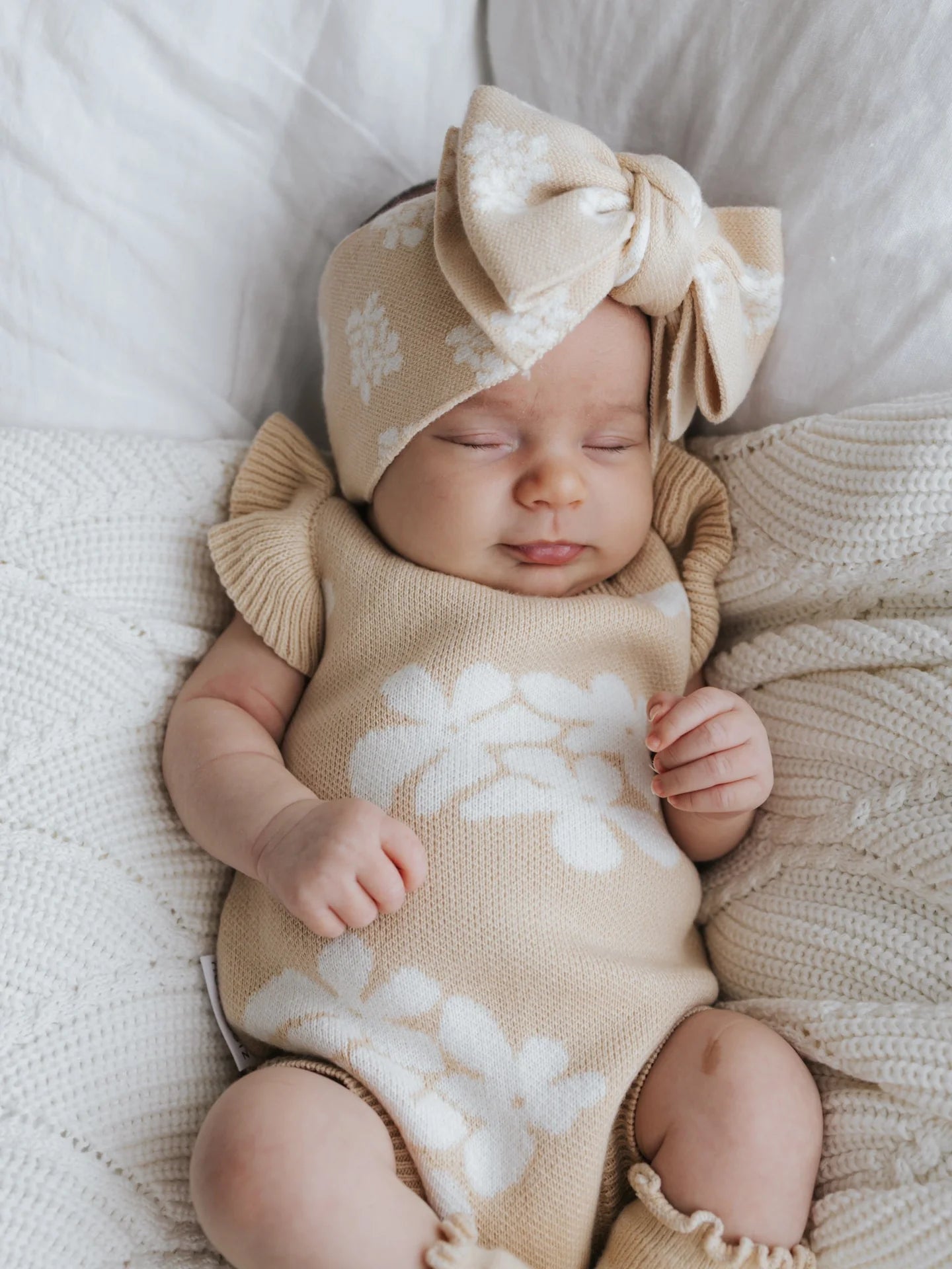 A baby is peacefully sleeping in the Frill Romper Banksy, a cream-colored knitted outfit adorned with white floral patterns designed by ZIGGY LOU. The romper features functional snaps for easy changing and comes with a matching headband accented by a large bow. The baby is lying on a soft, white textured blanket.