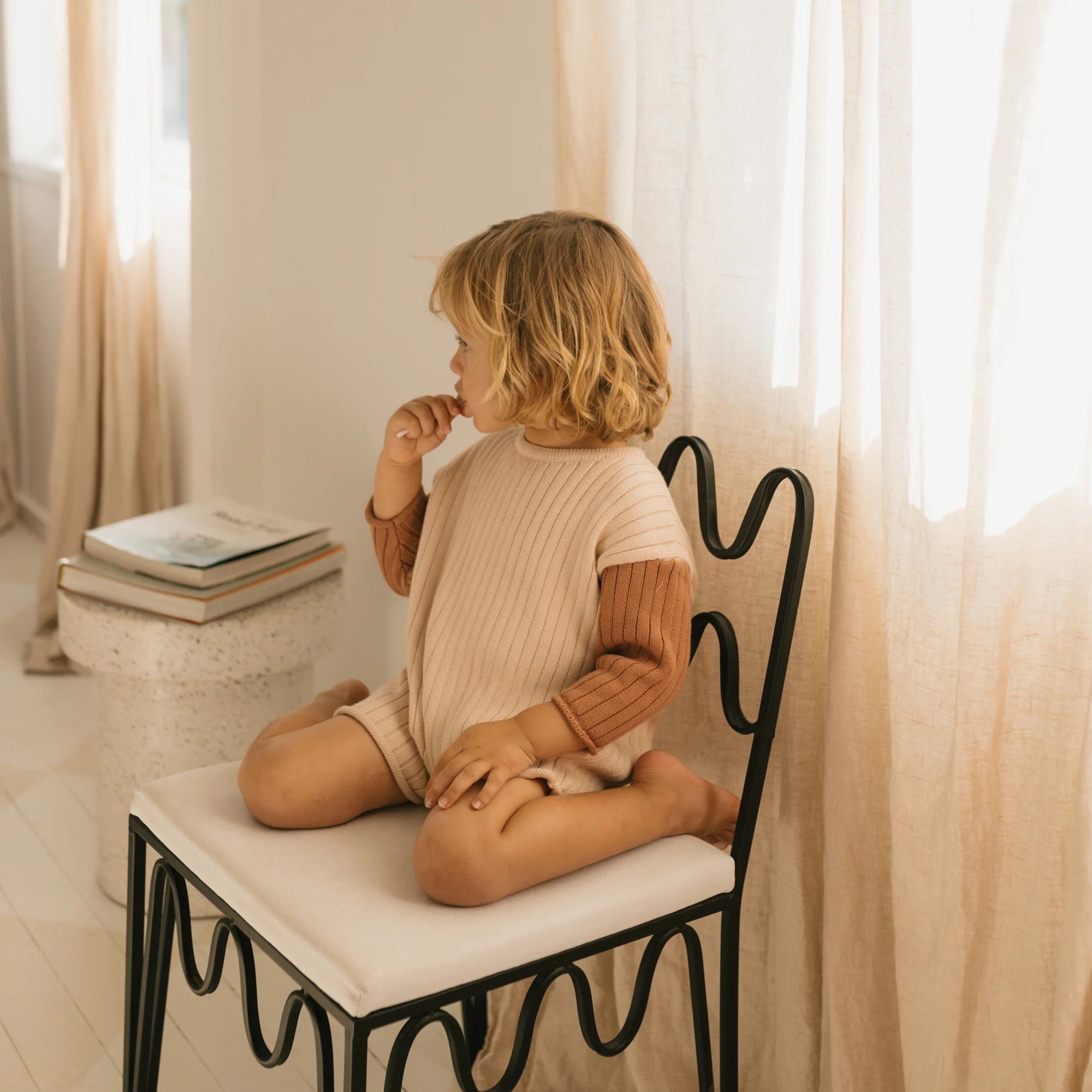 A young child with blond hair sits cross-legged on a decorative chair, clad in the Golden Knit Romper Caramel Sundae from GOLDEN CHILDREN. The light cream outfit with brown sleeves is crafted from soft cotton knit rib. The child gazes to the side, holding one finger near their mouth, as sunlight filters through light-colored curtains in the background.