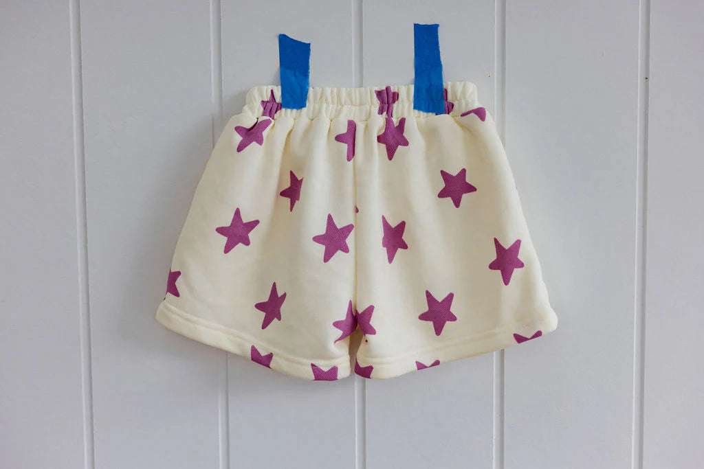 SUNDAY SIBLINGS' Kiddo Shorts Purple are crafted from cream french terry cotton, featuring a charming purple star print and displayed on a white wall with blue tape.