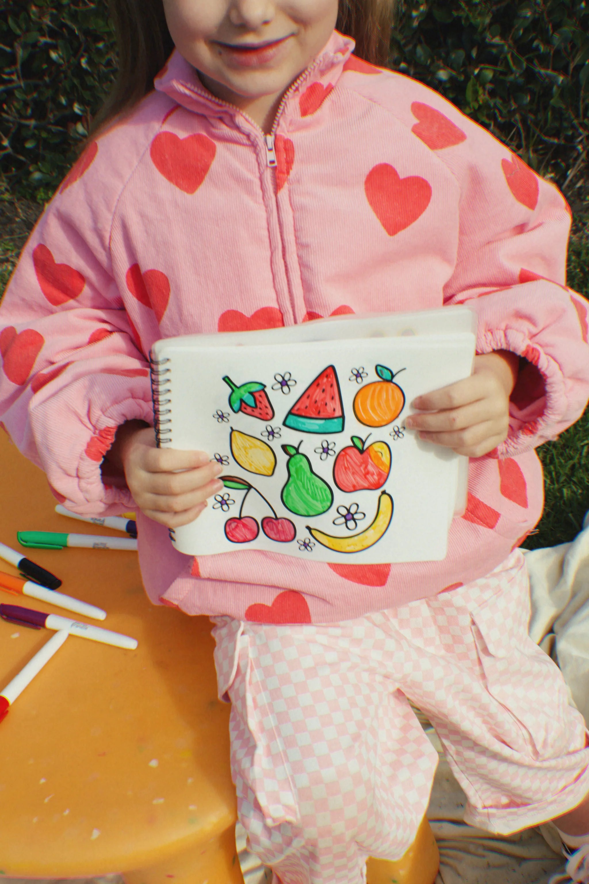 A child in a pink jacket with red hearts proudly displays their artwork from the OH MY FRANKIE ~ Oh My Scribbles Reusable Colouring Book, featuring vibrant fruits like watermelon and strawberries. At the table, non-toxic markers are scattered around, sparking kids' creativity.